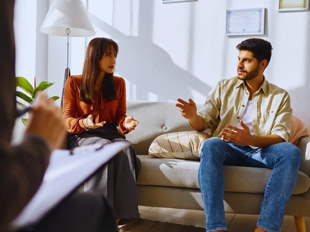 A woman and a man having a discussion in a therapy or counseling session, with a therapist taking notes in the foreground.
