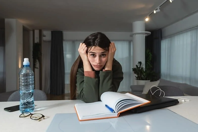 A young woman sitting at a table with a stressed or overwhelmed expression, resting her head in her hands, with a water bottle, glasses, open notebook, and a closed laptop in front of her in a modern, well-lit room.