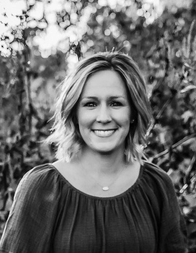 A woman with shoulder-length wavy hair smiling at the camera, outdoors with trees and foliage in the background.
