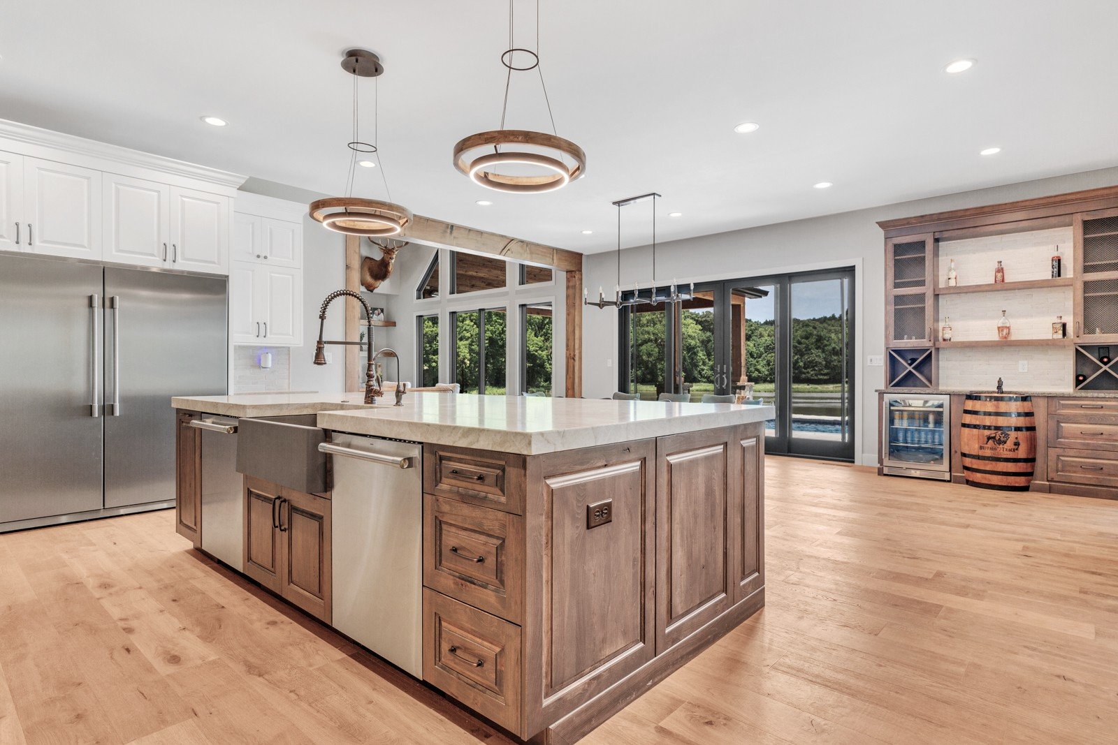 Modern kitchen with island, stainless steel refrigerator, and wooden cabinetry, overlooking outdoor landscape through large glass doors.