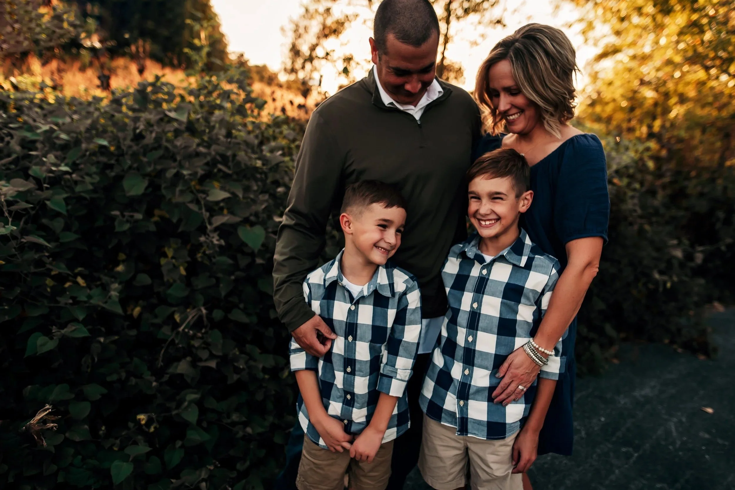 A family of four smiling and looking at each other outdoors during sunset, standing in front of a large bush with trees in the background.