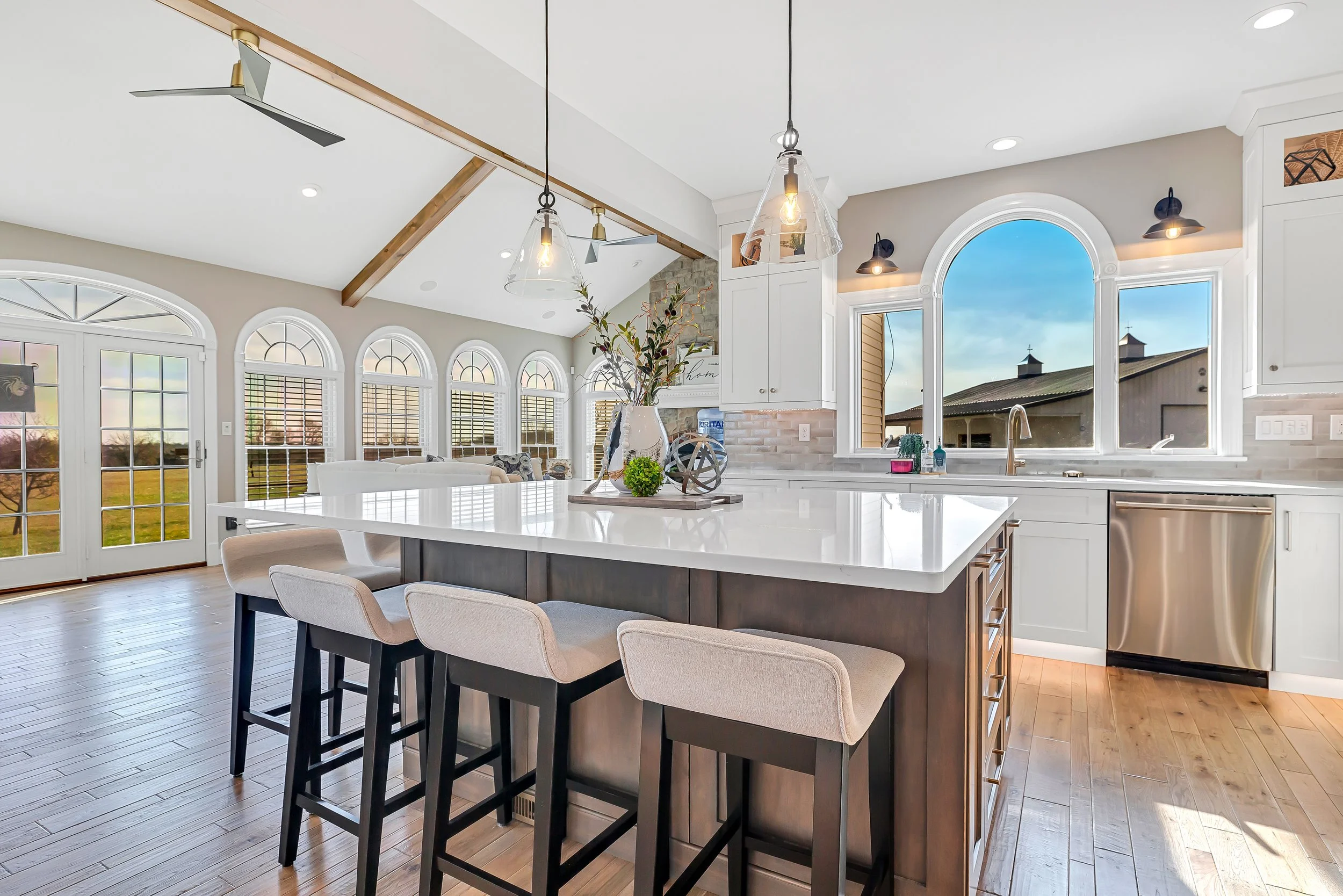 Bright, modern kitchen with a large island, white countertops, and pendant lights, featuring a view of a barn through an arched window.