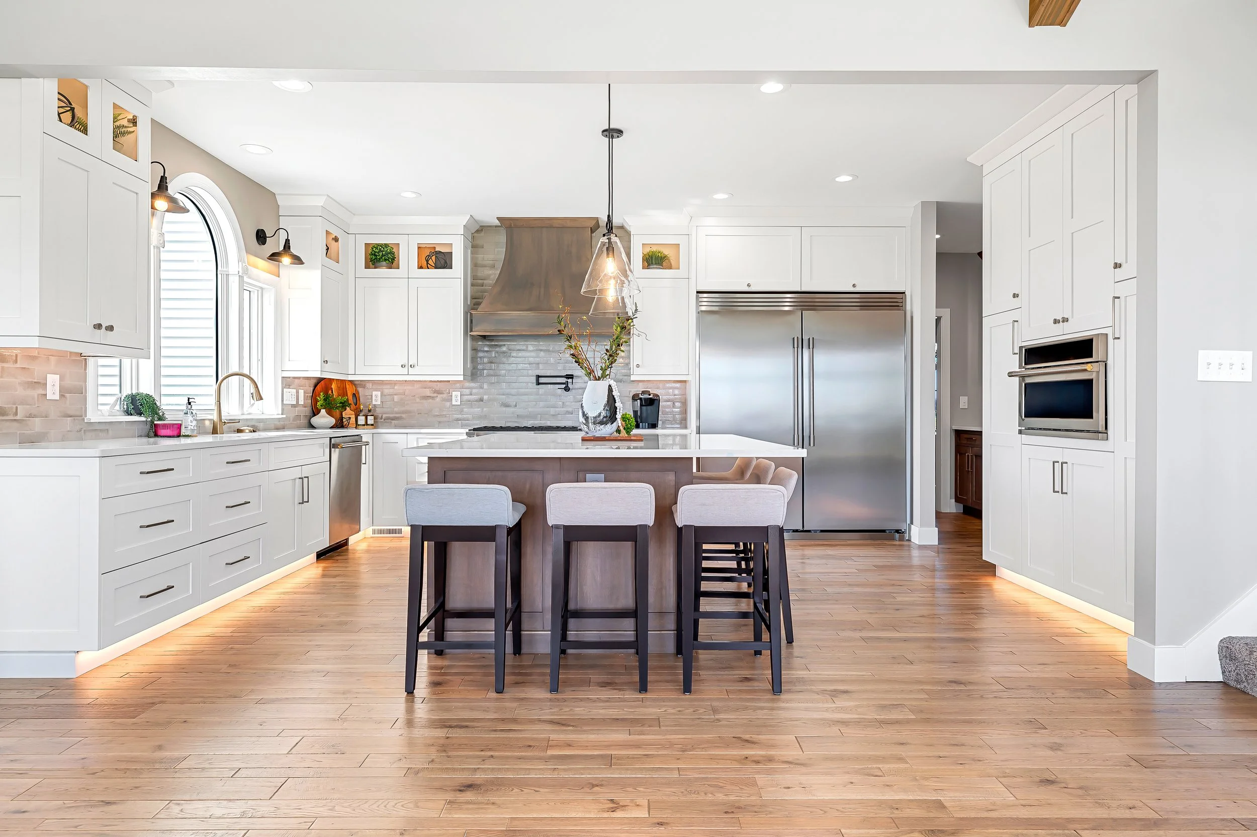 Modern kitchen with white cabinets, wooden flooring, stainless steel refrigerator, kitchen island with seating, window with blinds, decorative plants, and pendant lighting.