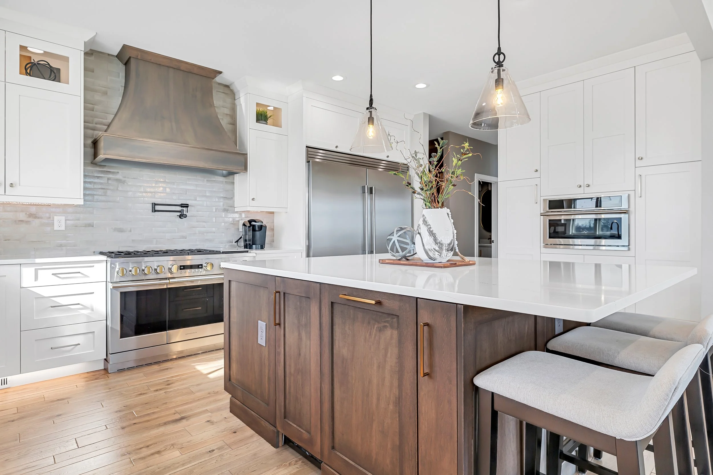 Modern kitchen with white cabinets, stainless steel appliances, a large island with wooden cabinets, pendant lights, and wooden flooring.