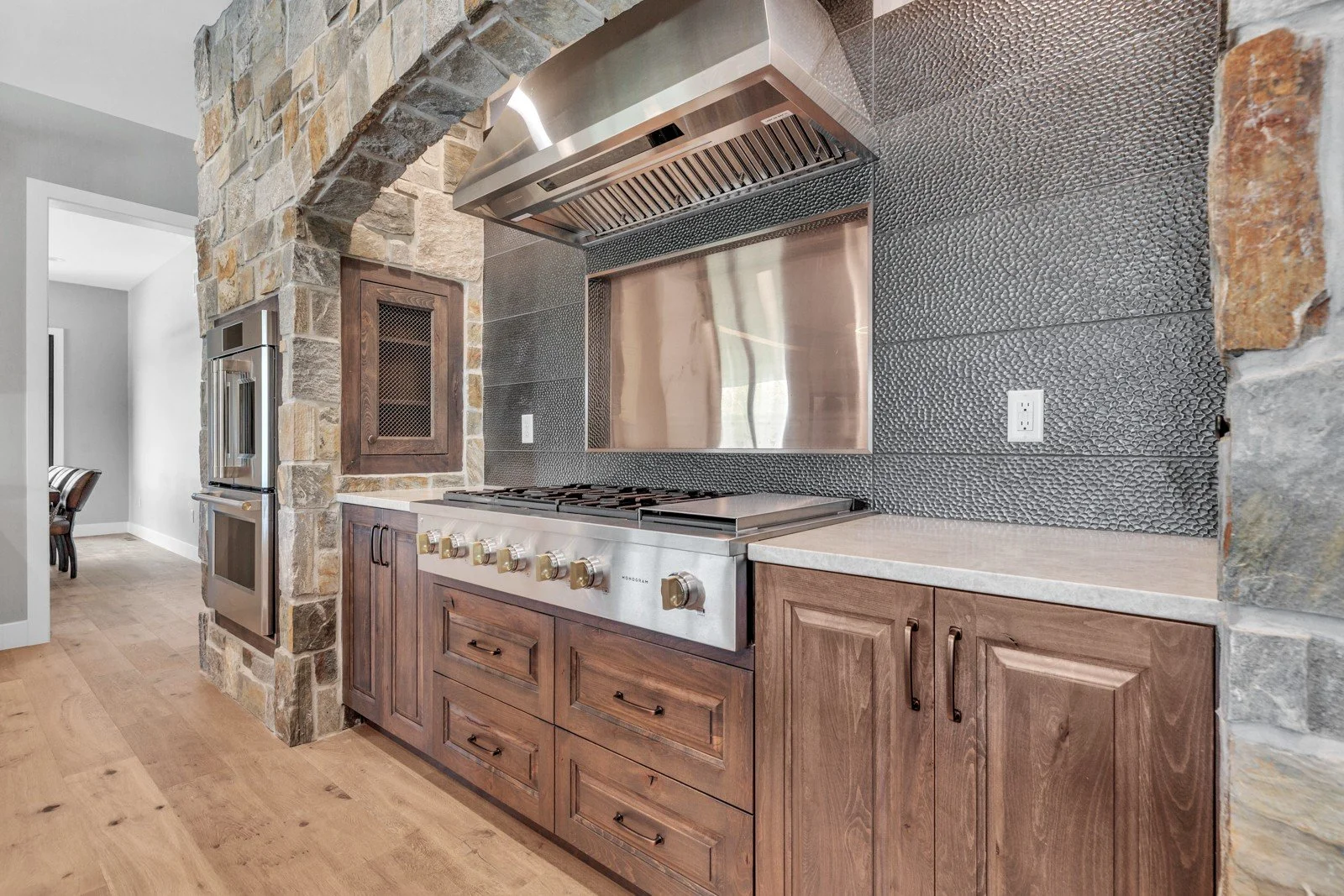 Modern kitchen with stainless steel stove, wooden cabinetry, stone accent wall, and black textured tile backsplash.