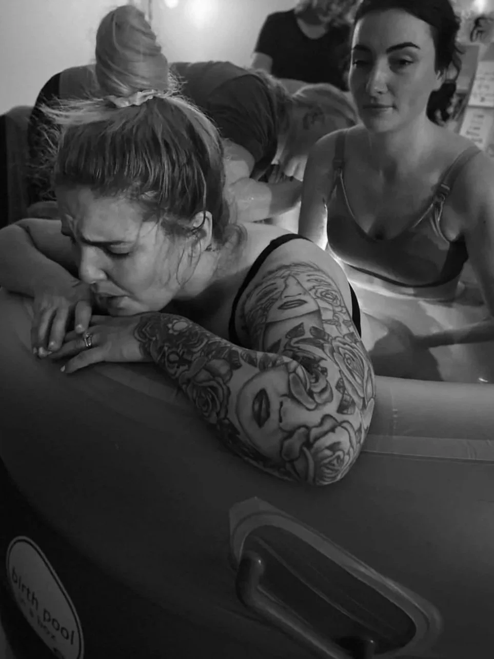 Black and white photo of a woman with tattoos on her arm, lying in a bathtub with her eyes closed. Two women are nearby, one with dark hair and the other with blond hair, both leaning over the tub.