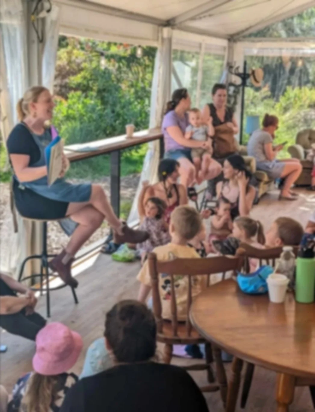 Group of women and children gathered indoors, some sitting on chairs and a stool, others on the floor, engaging in a casual gathering in a bright space with large windows and greenery outside.