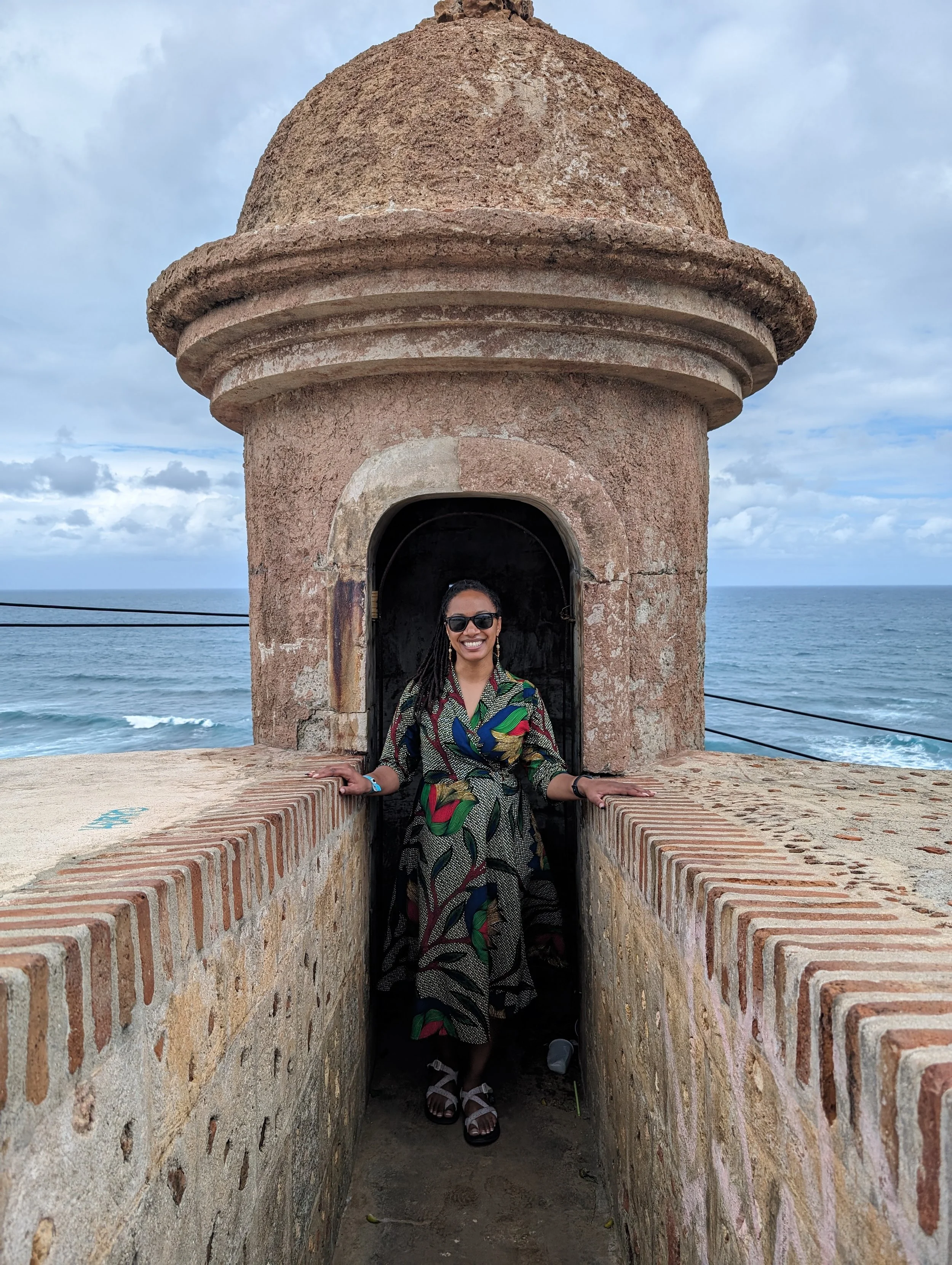 A woman with sunglasses, wearing a colorful patterned dress, standing in a small lookout tower on a cliff overlooking the ocean with cloudy skies.