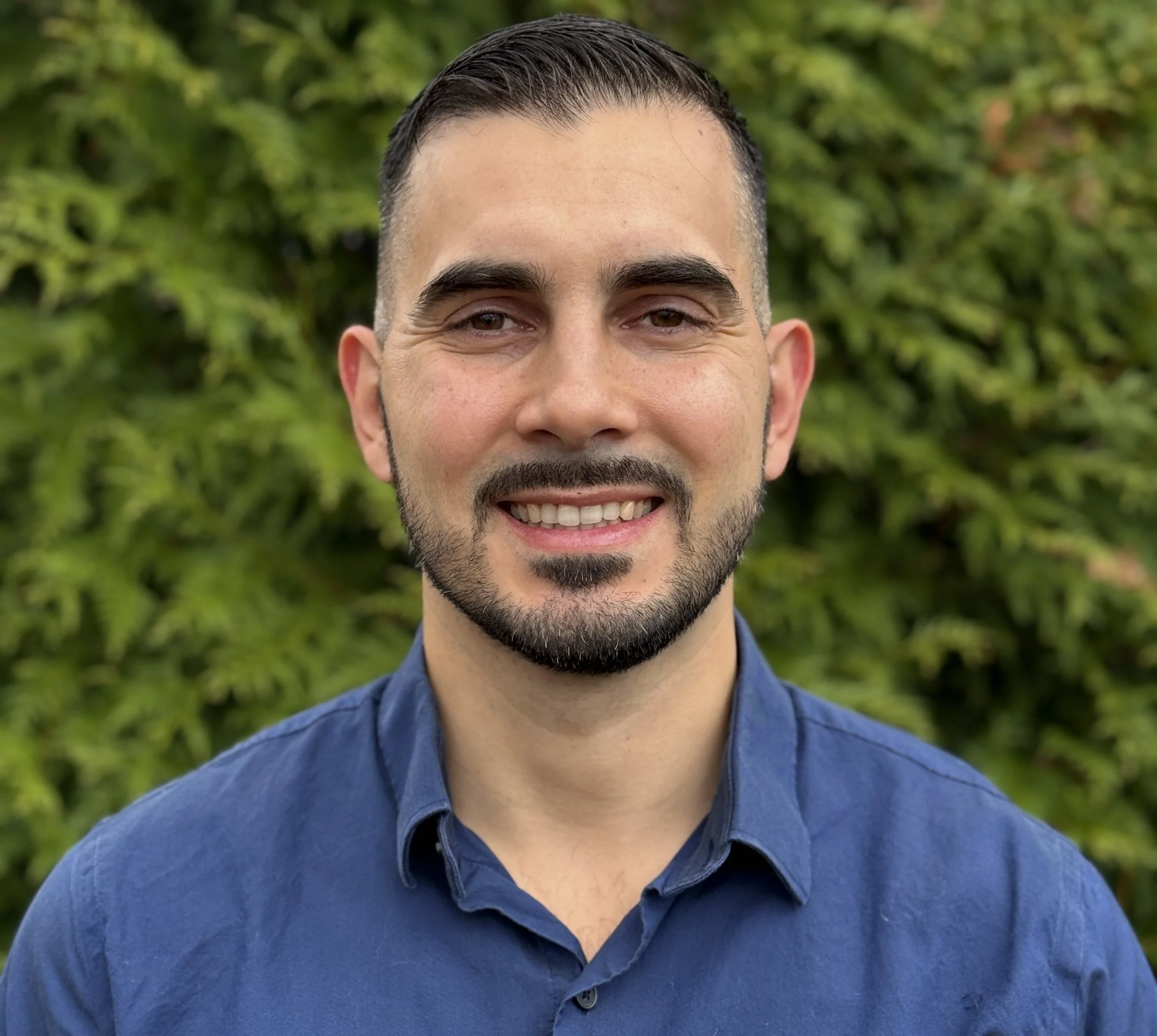 A smiling man with short dark hair, a trimmed beard and mustache, wearing a blue shirt, standing outdoors in front of green foliage.