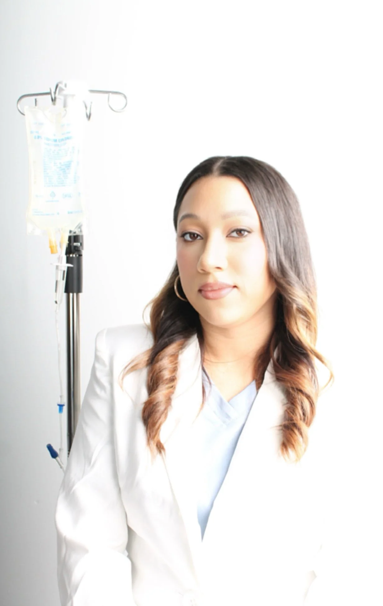 A woman with shoulder-length wavy hair, wearing a light-colored blazer and hoop earrings, sitting next to an IV stand with a medical infusion bag, in a bright room.