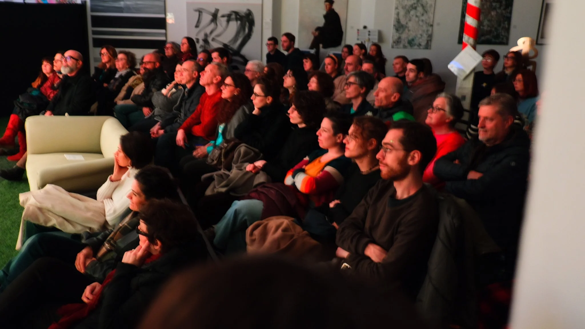Pubblico seduto in sala che guarda un evento teatrale a Lanarchico Prato, con arte moderna alle pareti.