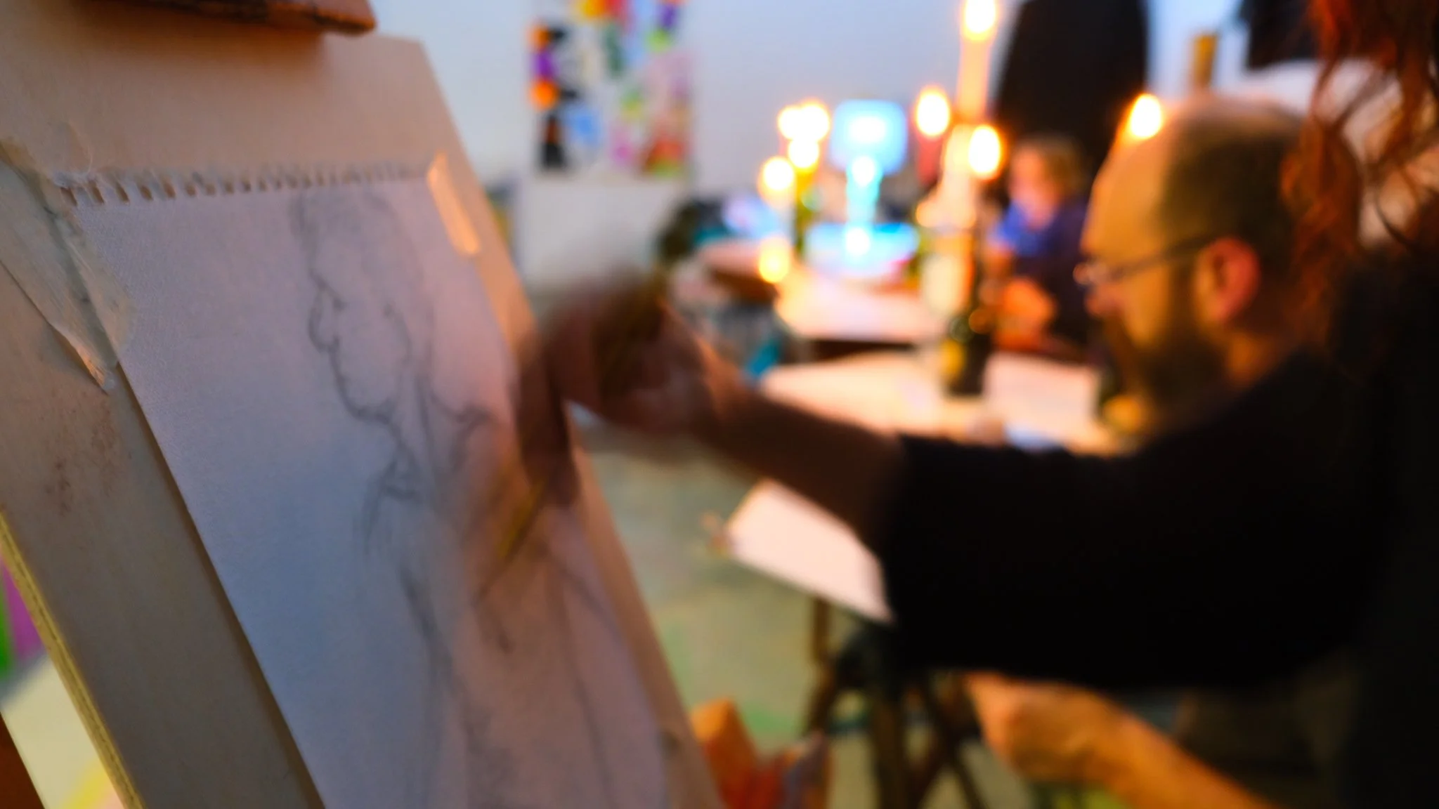 Artista che disegna un quadro su una tavoletta di legno, in un ambiente con luci calde e altri artisti al lavoro sullo sfondo. Disegno nudo dal vero organizzato a Prato, in Toscana, nel centro di arte contemporanea Lanarchico