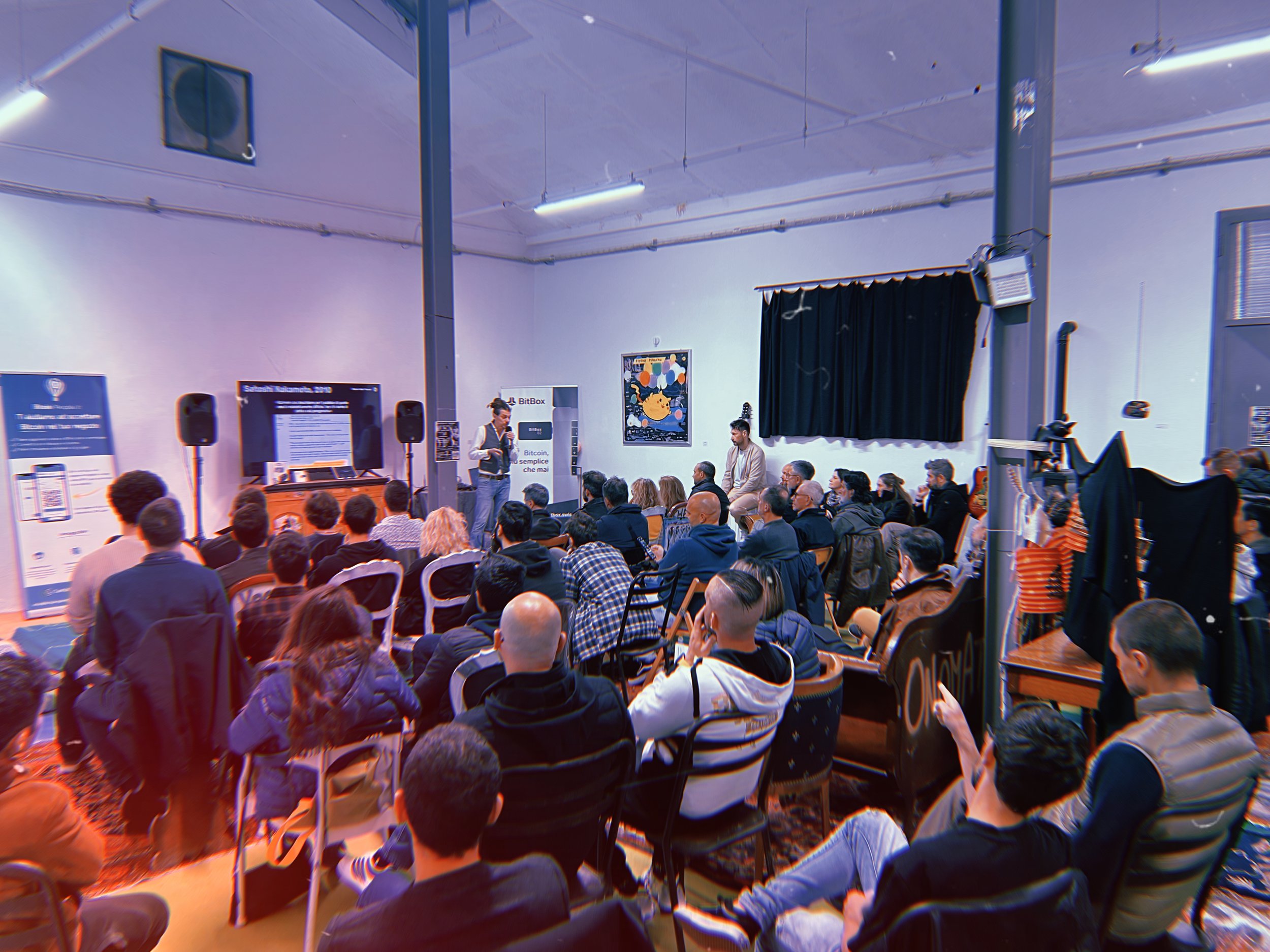 Una sala piena di persone che assistono a una conferenza di divulgazione a tema Bitcoin. Lanarchico si occupa anche di formazione e seminari, le aziende possono affittare lo spazio per coinvolgere un pubblico più cool 