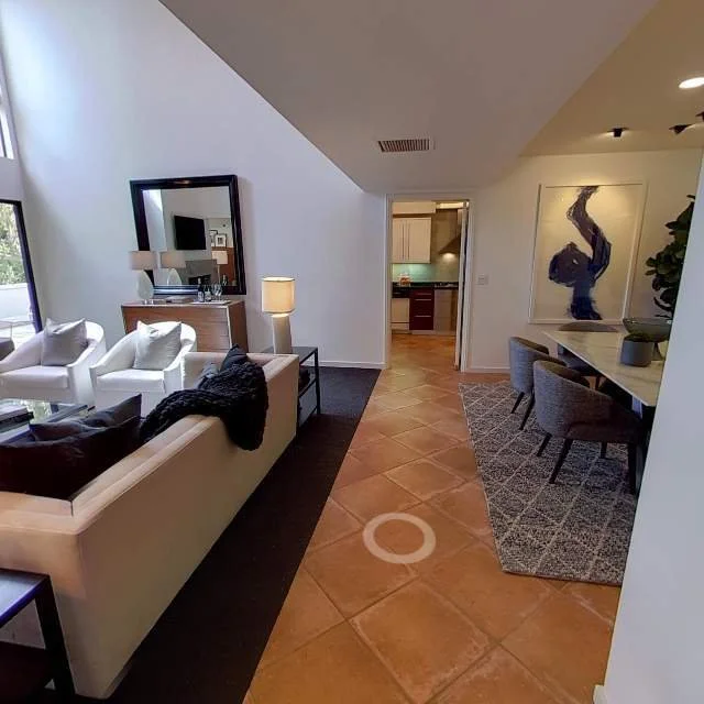 Living room with white sofa, two white armchairs, lamps, a large mirror, and artwork of an elephant on the wall; adjacent dining area with gray chairs and a rug, kitchen visible in the background.