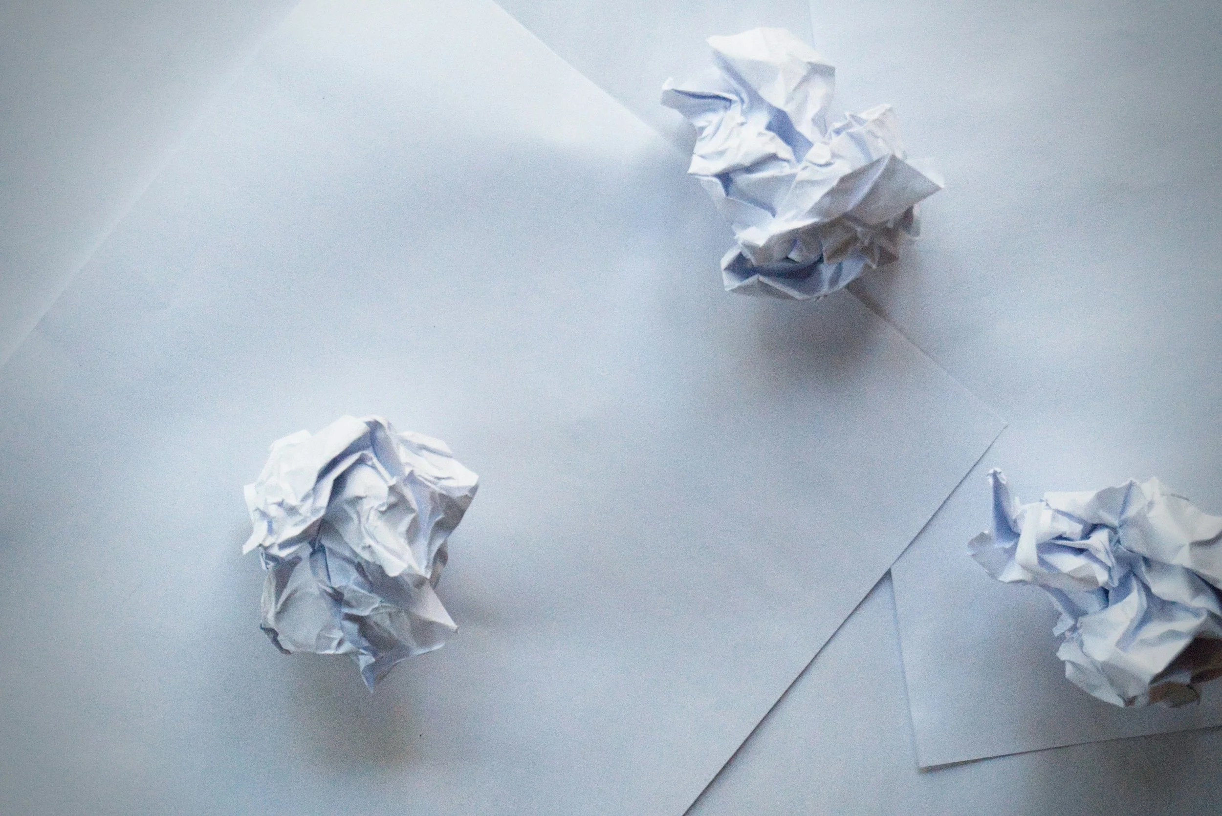 Three crumpled sheets of white paper on a white surface with a sheet of paper underneath one of them.