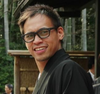 A man with short brown hair, wearing glasses and a dark jacket, smiling outdoors in front of a wooden structure and trees.