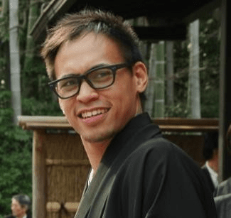 A young man with glasses, smiling outdoors, wearing a black jacket, and standing near a wooden structure with trees in the background.