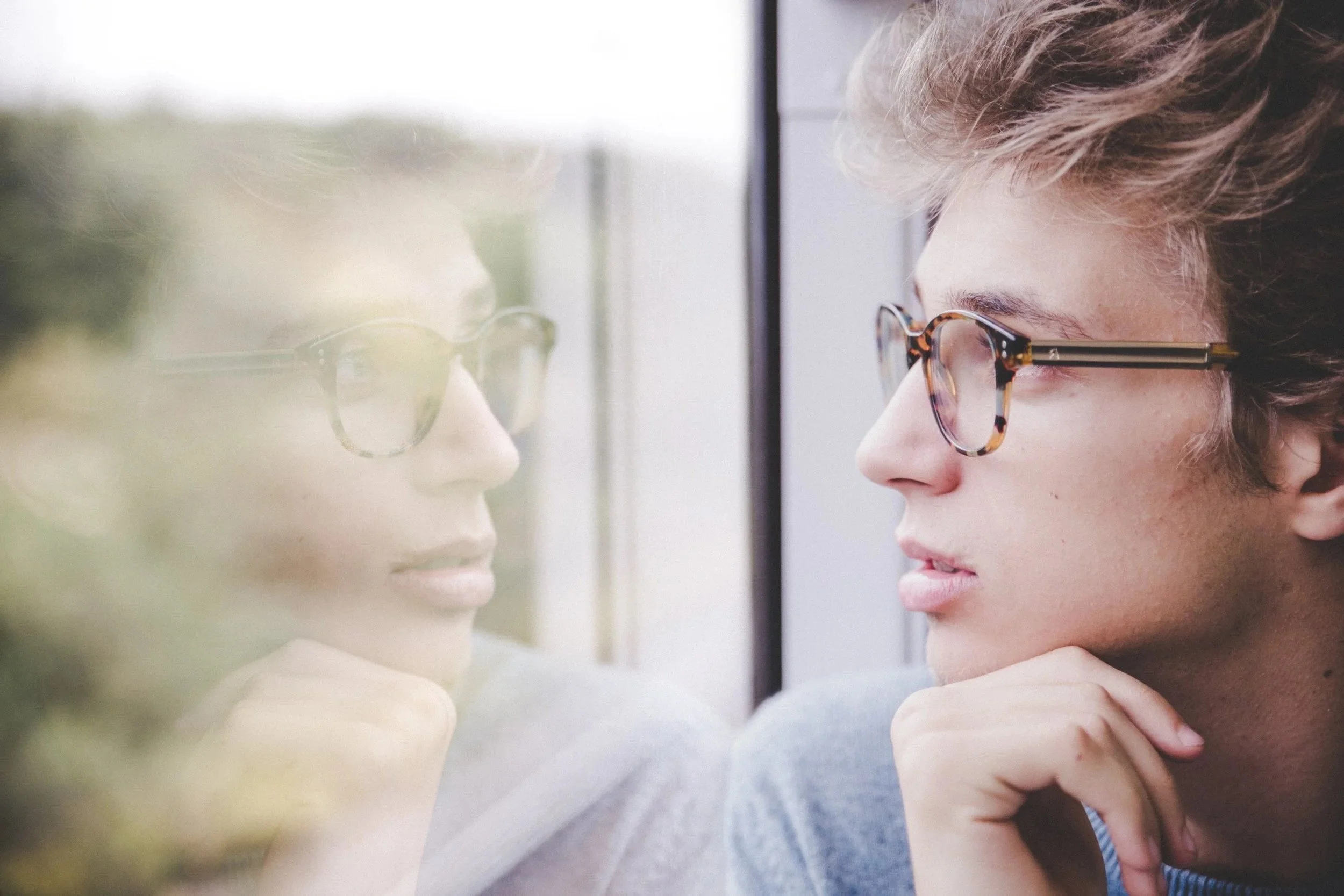 Young man with glasses looking at his reflection on a window.