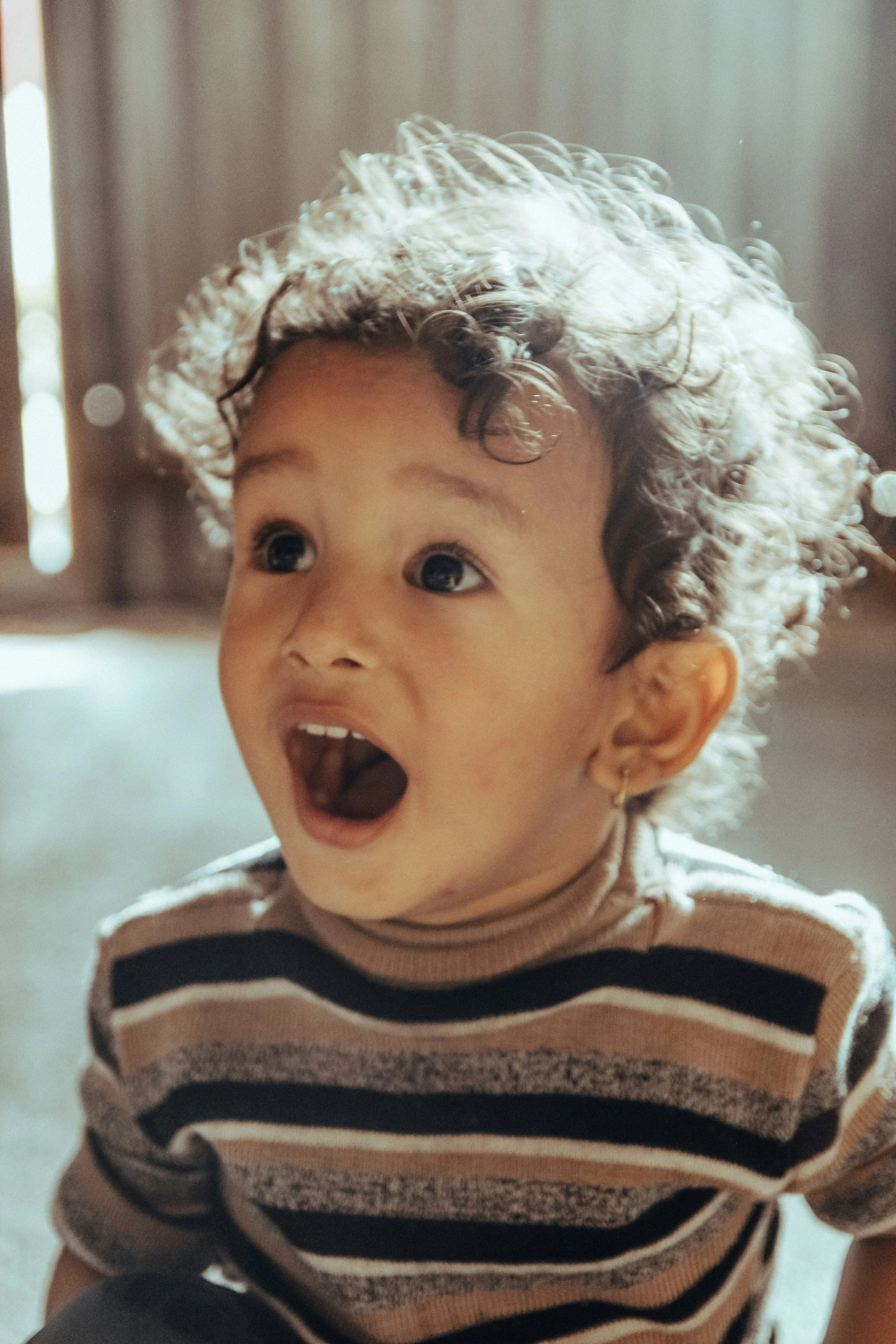 A young child with curly blonde hair, wearing a striped shirt, with an excited expression and mouth open.