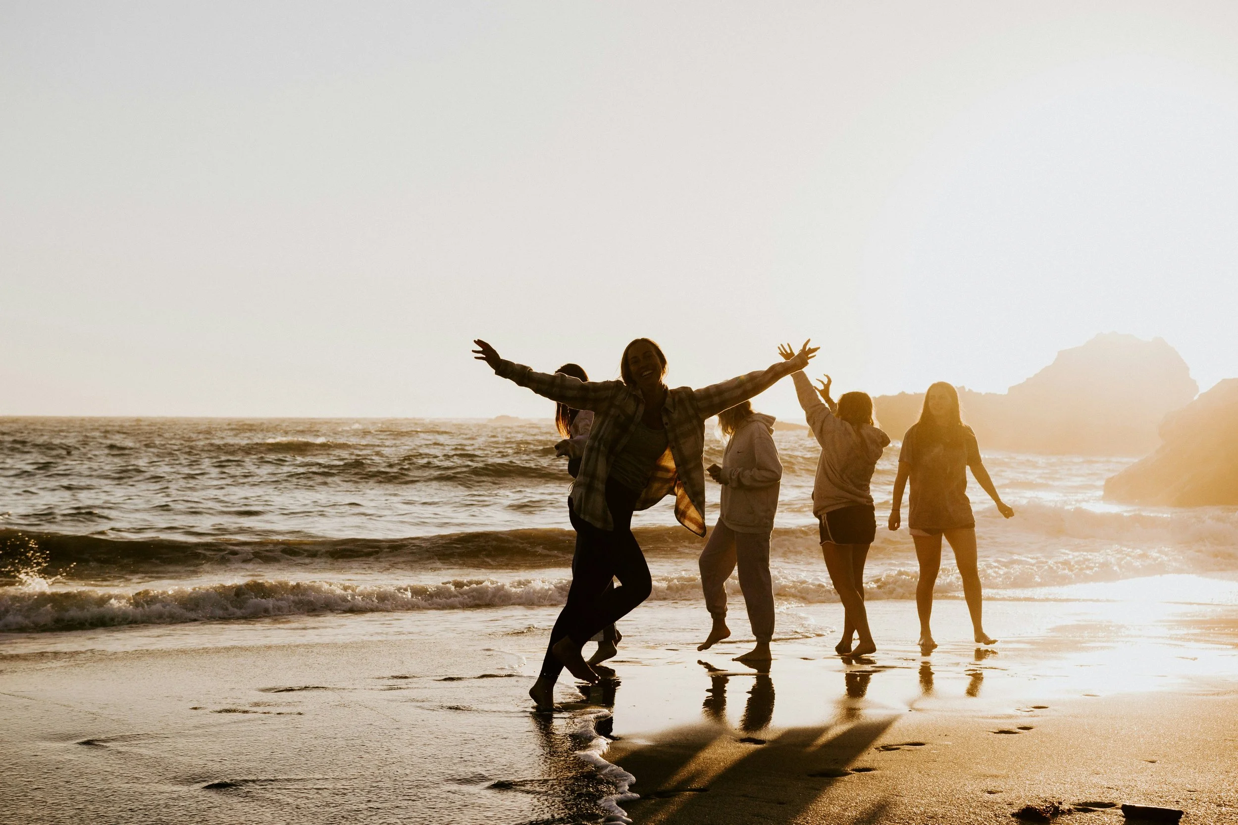 A group of five people on the beach during sunset, smiling and playing near the water. One person is jumping with arms outstretched, and the others are walking or standing along the shoreline.