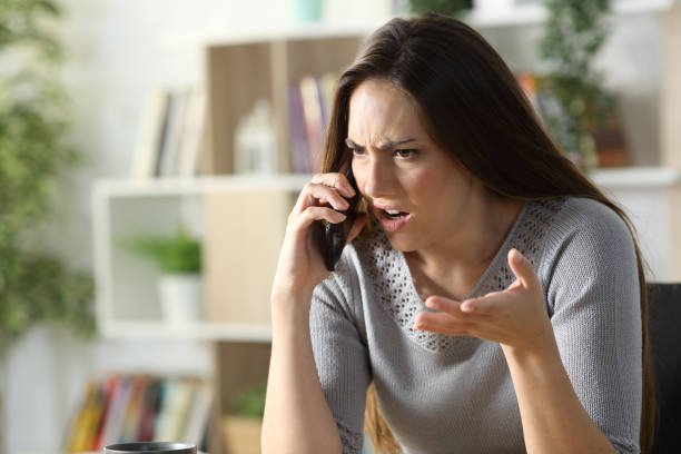 Women with long brown hair talking on the phone, looking confused or upset, indoors with bookshelves in the background.