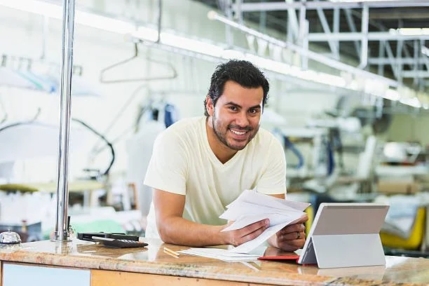 A man smiling and holding papers in a workshop or workspace. There is a tablet or laptop on the counter in front of him.
