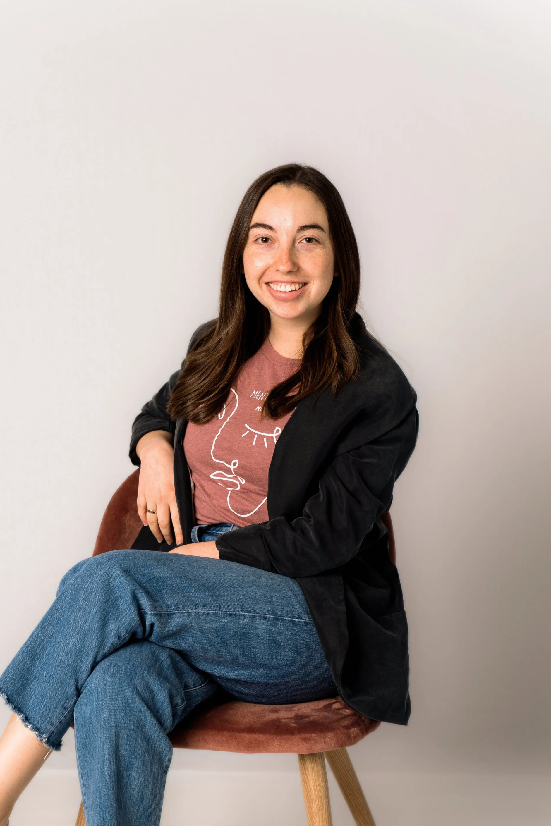 A woman with dark brown hair and fair skin sitting on a brown chair against a white wall, smiling, wearing a black jacket, a mauve t-shirt with a line drawing of a face, and blue jeans.