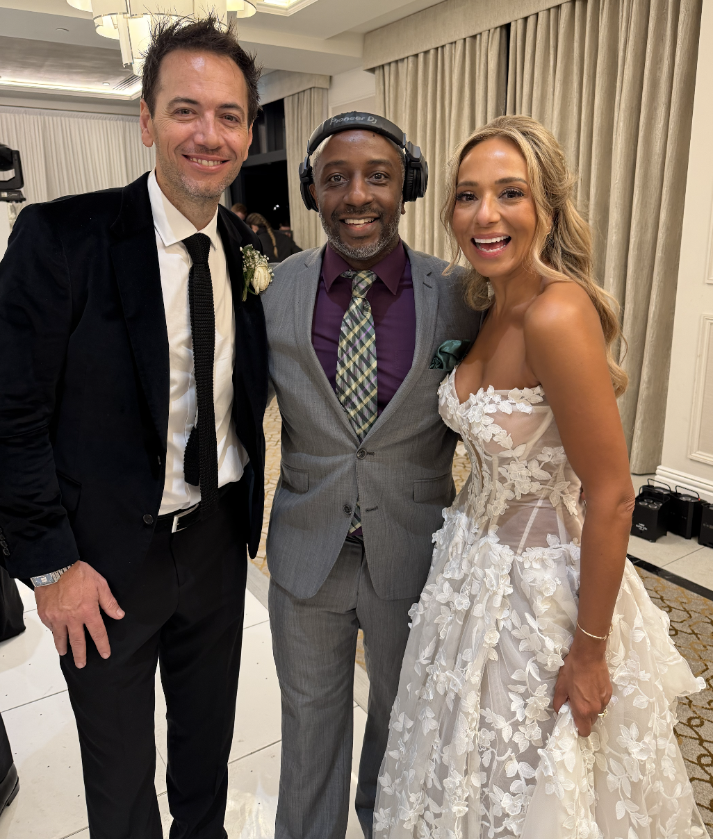 A groom in a black suit and white shirt standing next to his newly wedded wife in a white wedding dress, both smiling. They are standing next to DJ Mr Drew at their wedding reception.