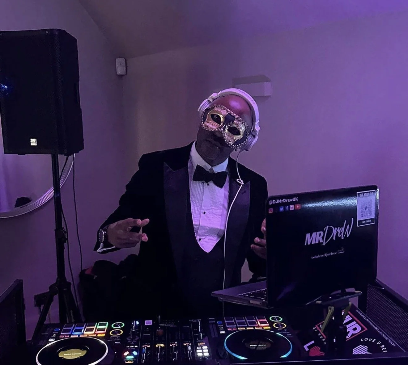 DJ Mr Drew wearing a masquerade mask, headphones, and a tuxedo, standing behind DJ equipment in a dimly lit room.