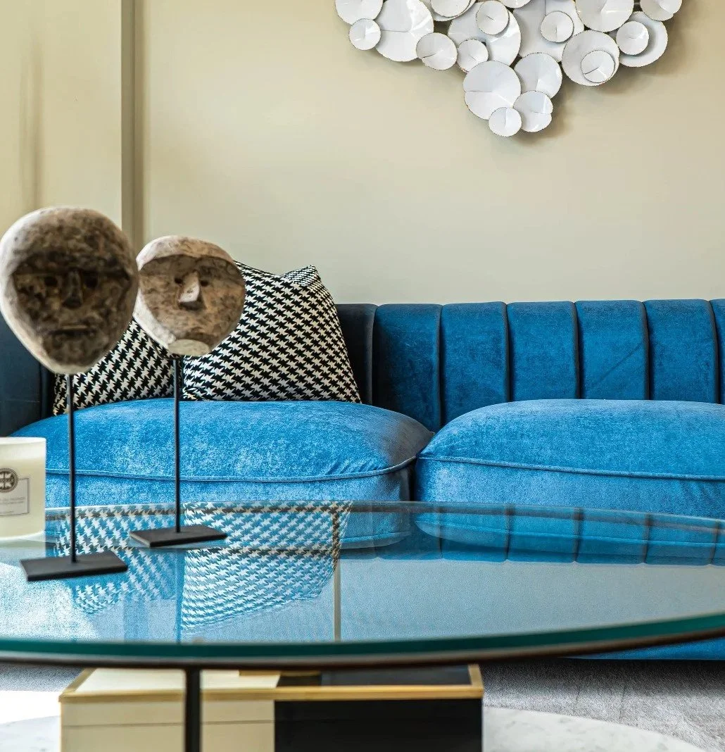 Living room with a blue velvet sofa, black and white houndstooth pillows, decorative wood sculptures on stands, a glass coffee table, and abstract white wall art.
