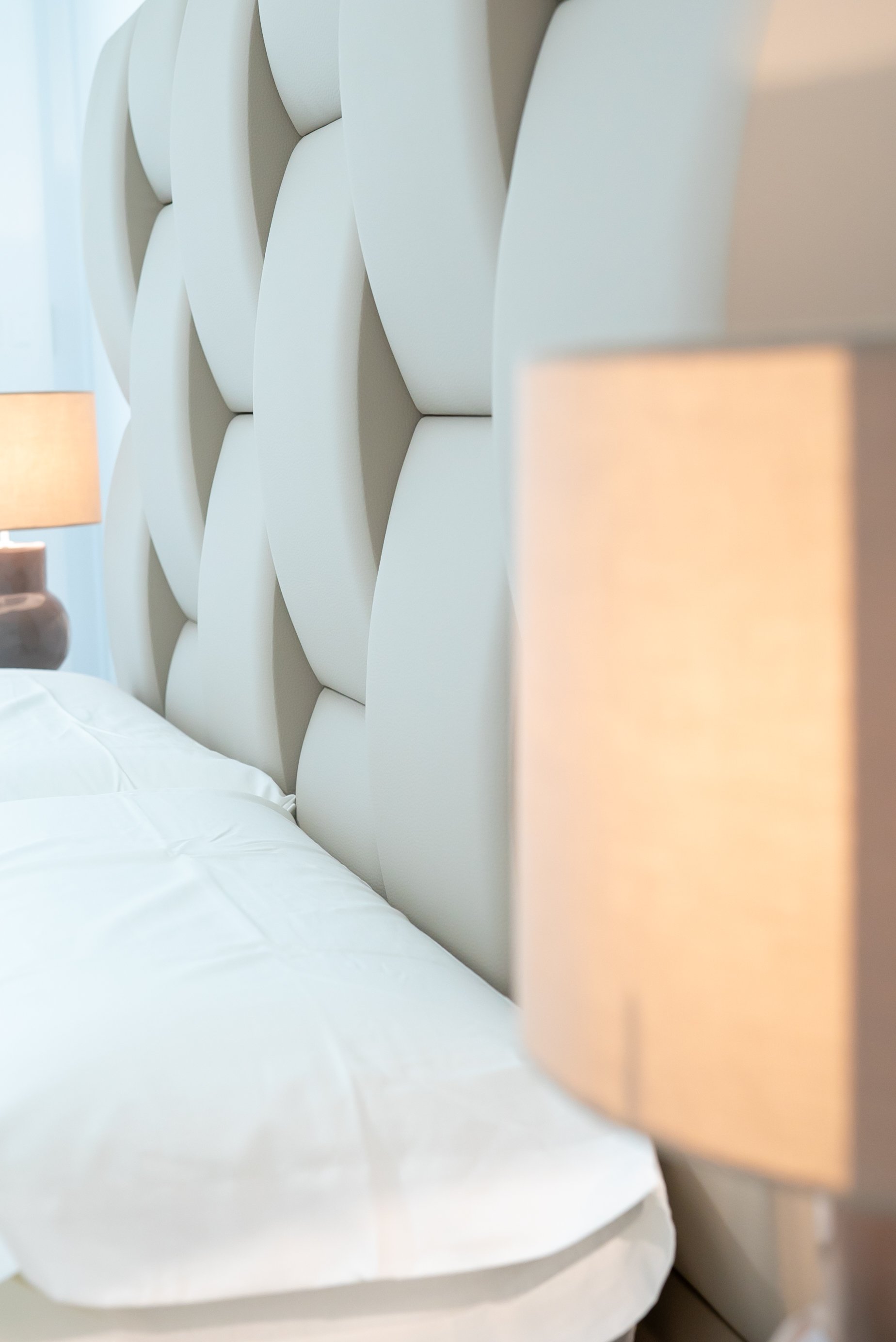 Close-up of a modern, white, padded headboard on a bed with neatly made white bedding. A beige lampshade is partially visible on the left side.