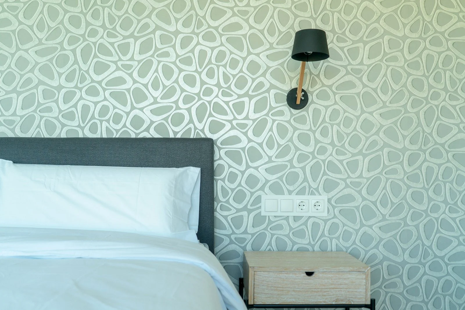 A bedroom with a white bed, a gray headboard, a small wooden nightstand, a black wall-mounted lamp, and a patterned wall with light gray and white abstract shapes. There are also electrical outlets on the wall.