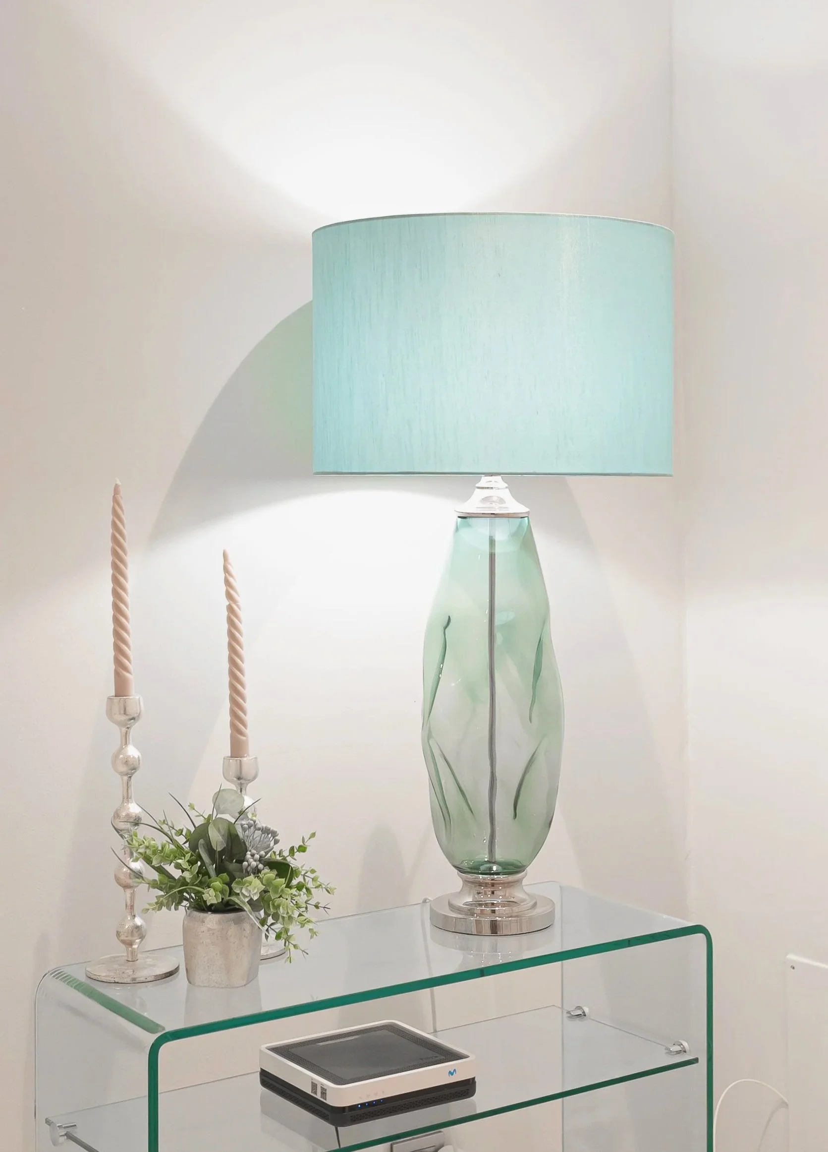 A decorative display with two tall candlesticks with twisted candles, a white vase with greenery, a glass table with a cake slicer, a large green glass vase with a lamp shade on it, and a small floral arrangement on a glass table.
