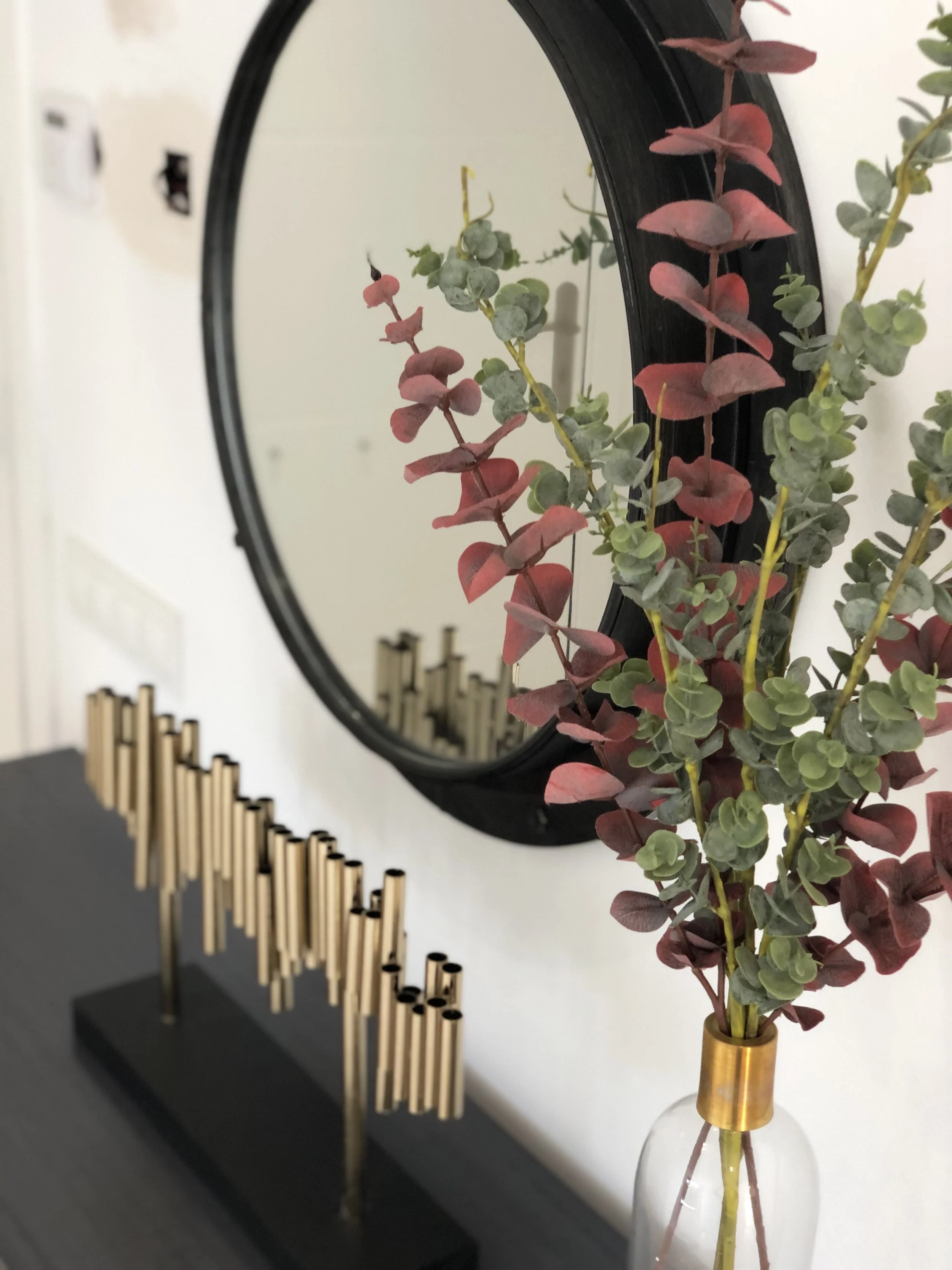 Decorative mirror reflecting green and red foliage in a tall glass vase with a gold band, placed on a black surface beside a black object with gold chopsticks.
