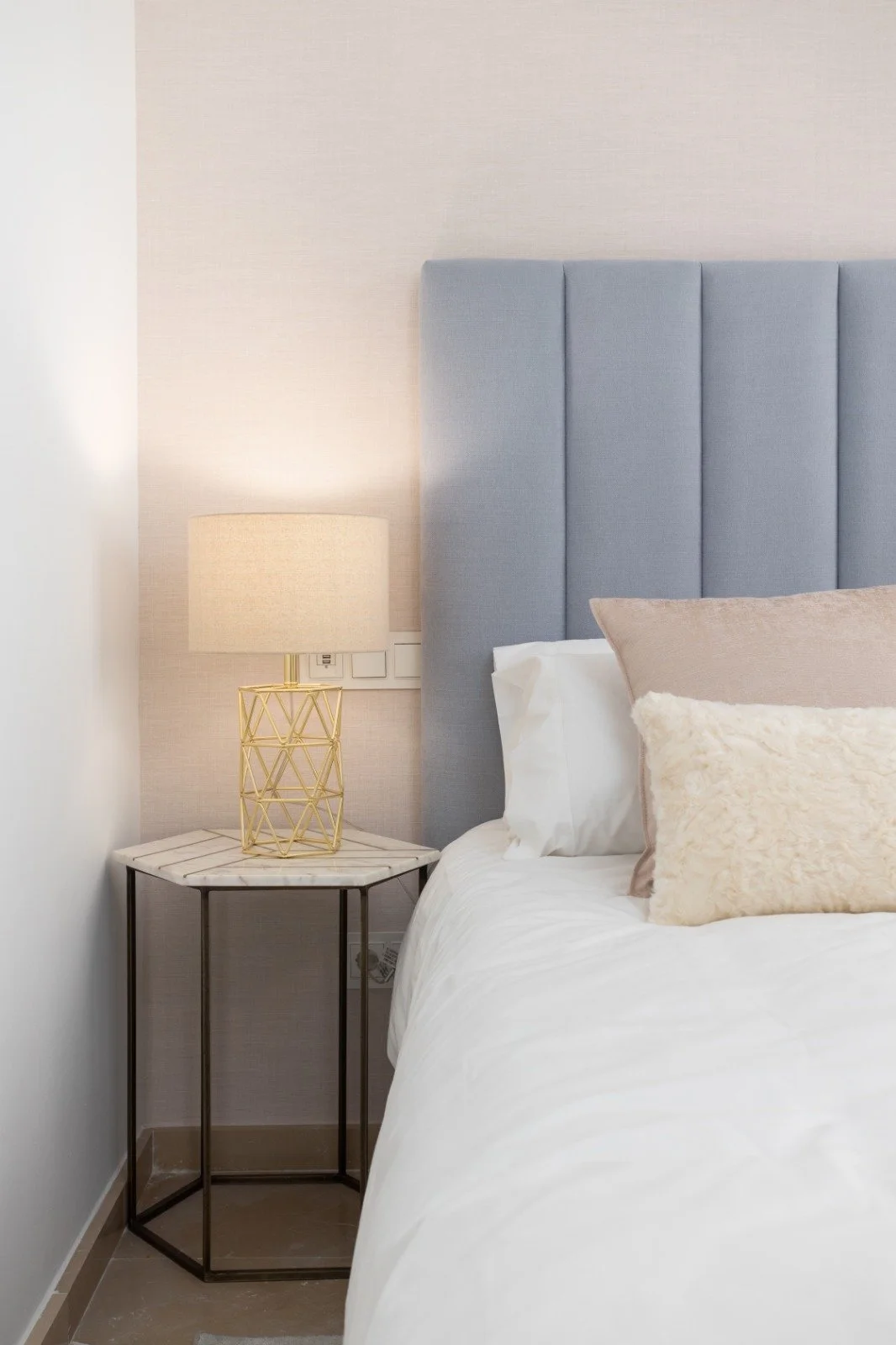 Close-up of a bedroom nightstand with a geometric gold lamp, beside a bed with a light blue upholstered headboard and beige and white pillows.