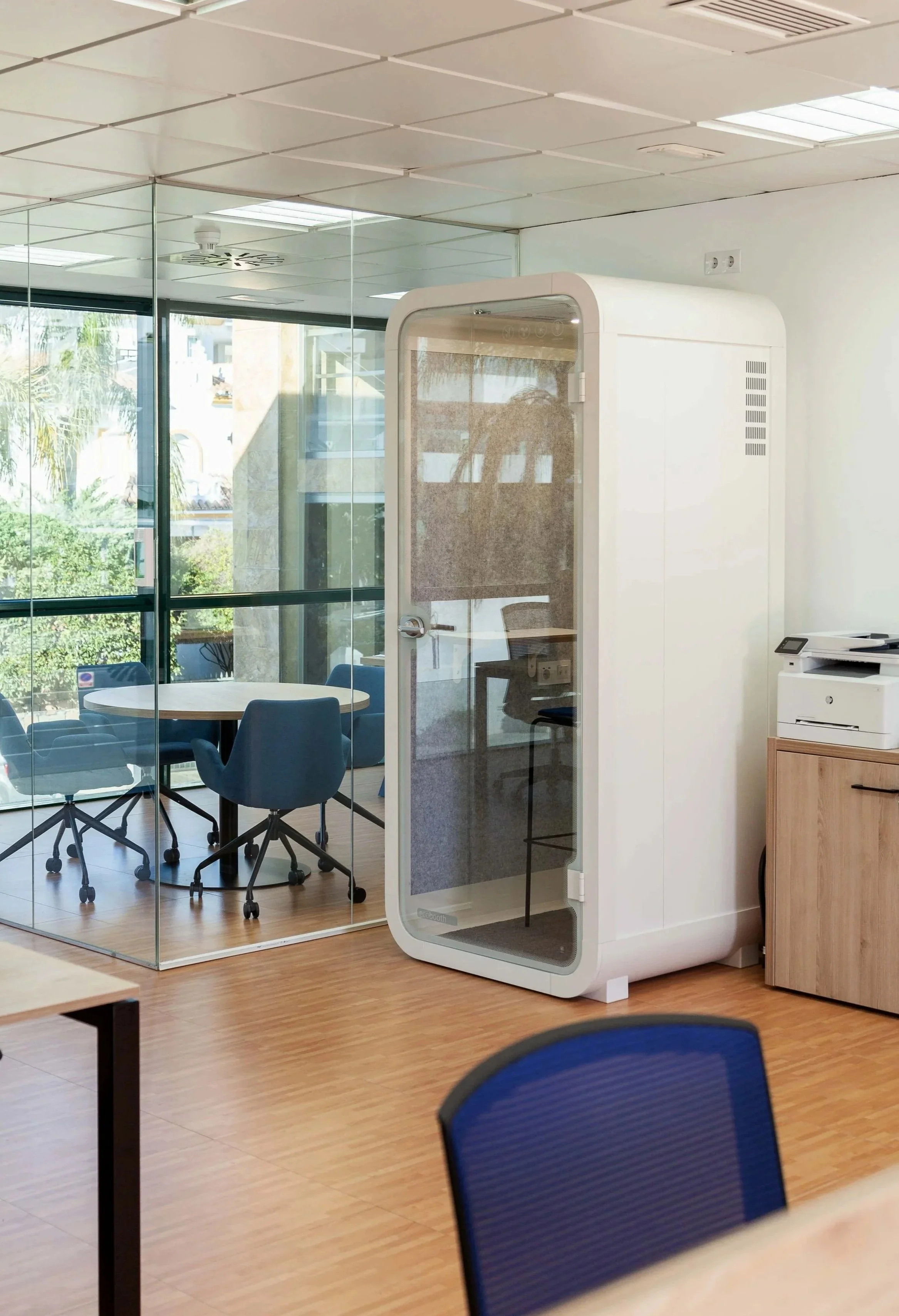An office conference room with glass walls, a round table, several blue office chairs, and a white enclosed phone booth. There is a window with a view of trees and buildings outside, and nearby includes a wooden cabinet and a printer.