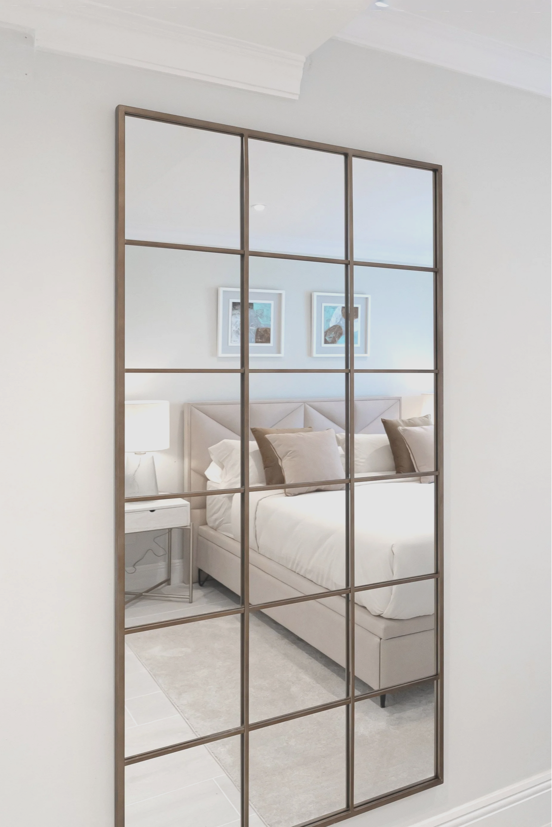 A large room with a mirrored wall reflecting a bedroom with a white bed, beige headboard, beige pillows, a white nightstand, and framed artwork on the wall.