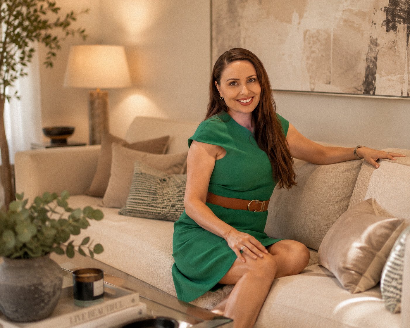 A woman with long brown hair is sitting on a beige sofa in a living room, smiling and looking at the camera. She is wearing a green dress with a brown belt, and has her left arm resting on the back of the sofa.