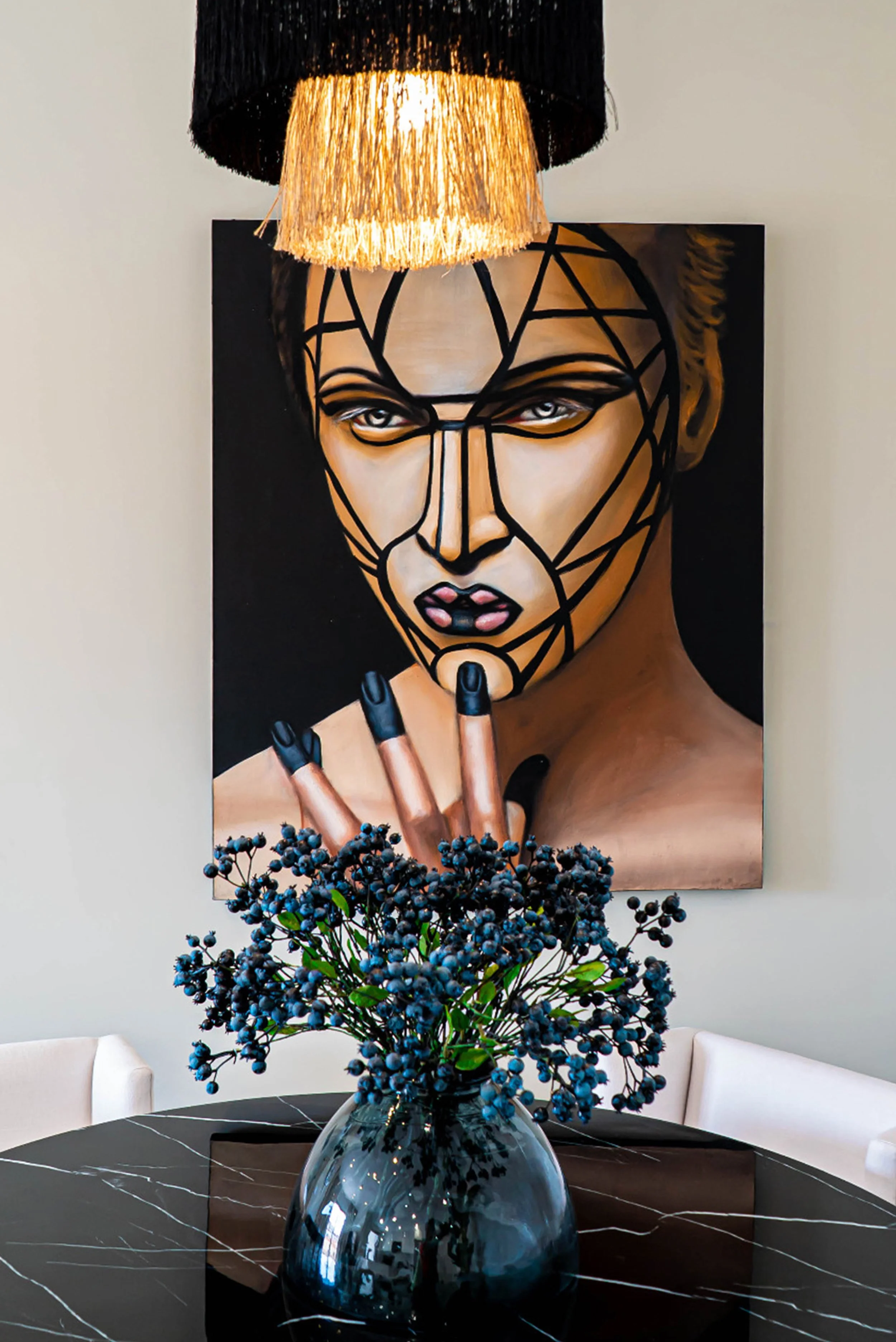 Colorful modern artwork of a woman with geometric facial features and piercing eyes, hanging on a beige wall behind a black round table with a dark blue vase filled with dark purple berries in the foreground.