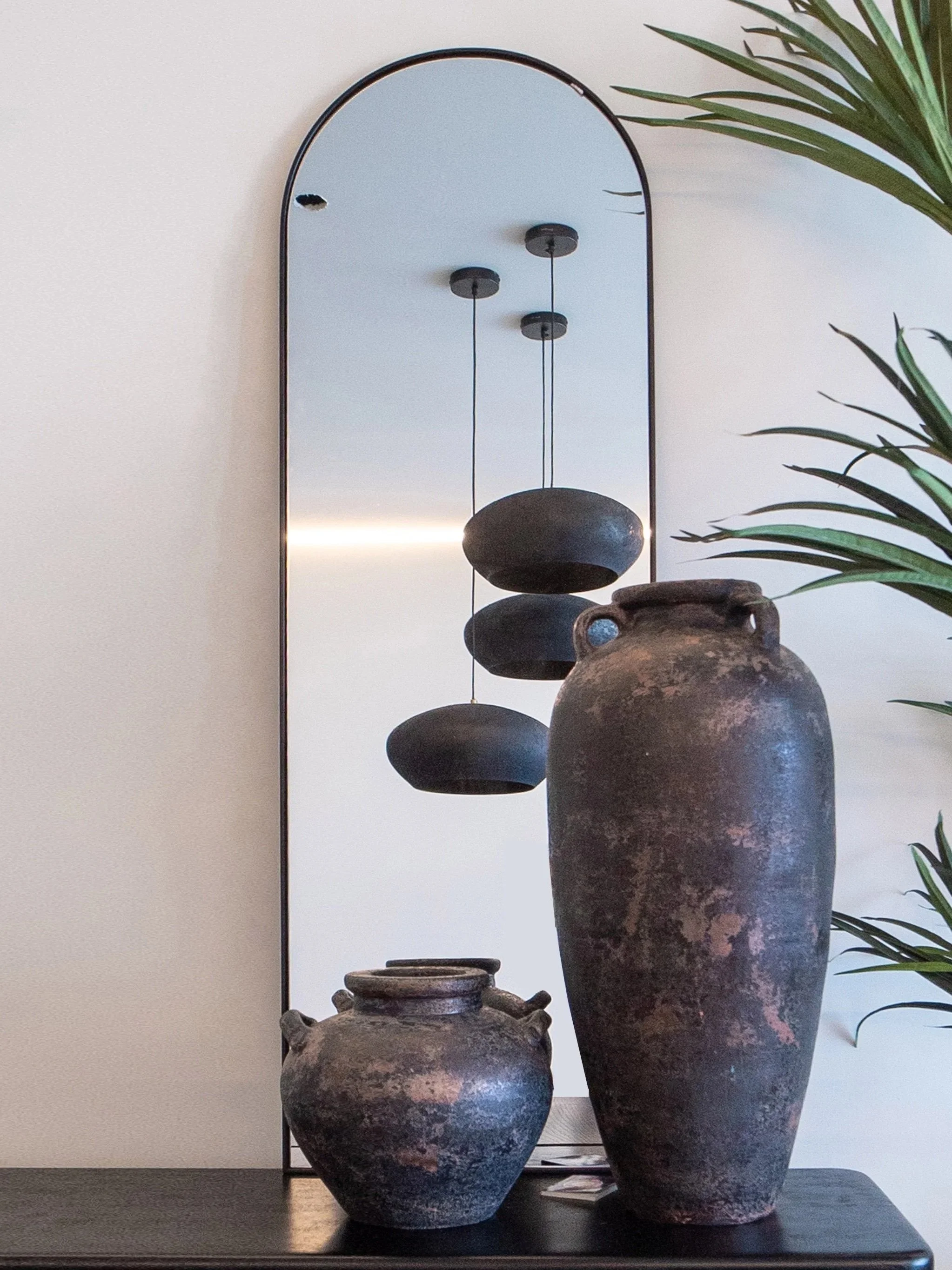A display of vintage pottery and vases on a black table, with a mirror in the background and a plant on the right.