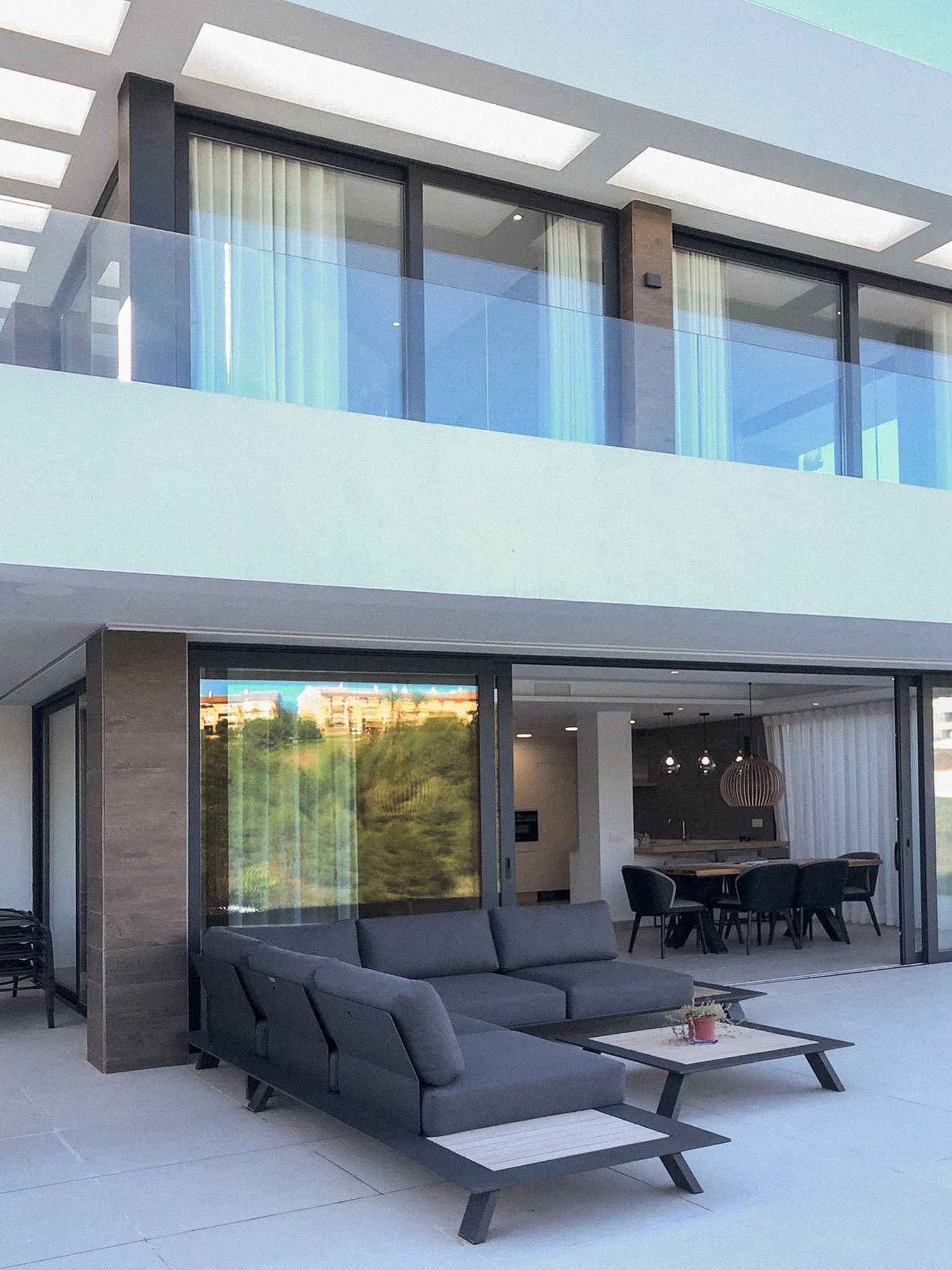 Living room with black couches, a coffee table with a small plant, open dining area with a round table, and a kitchen in the background. Large sliding glass doors lead to an outdoor view.