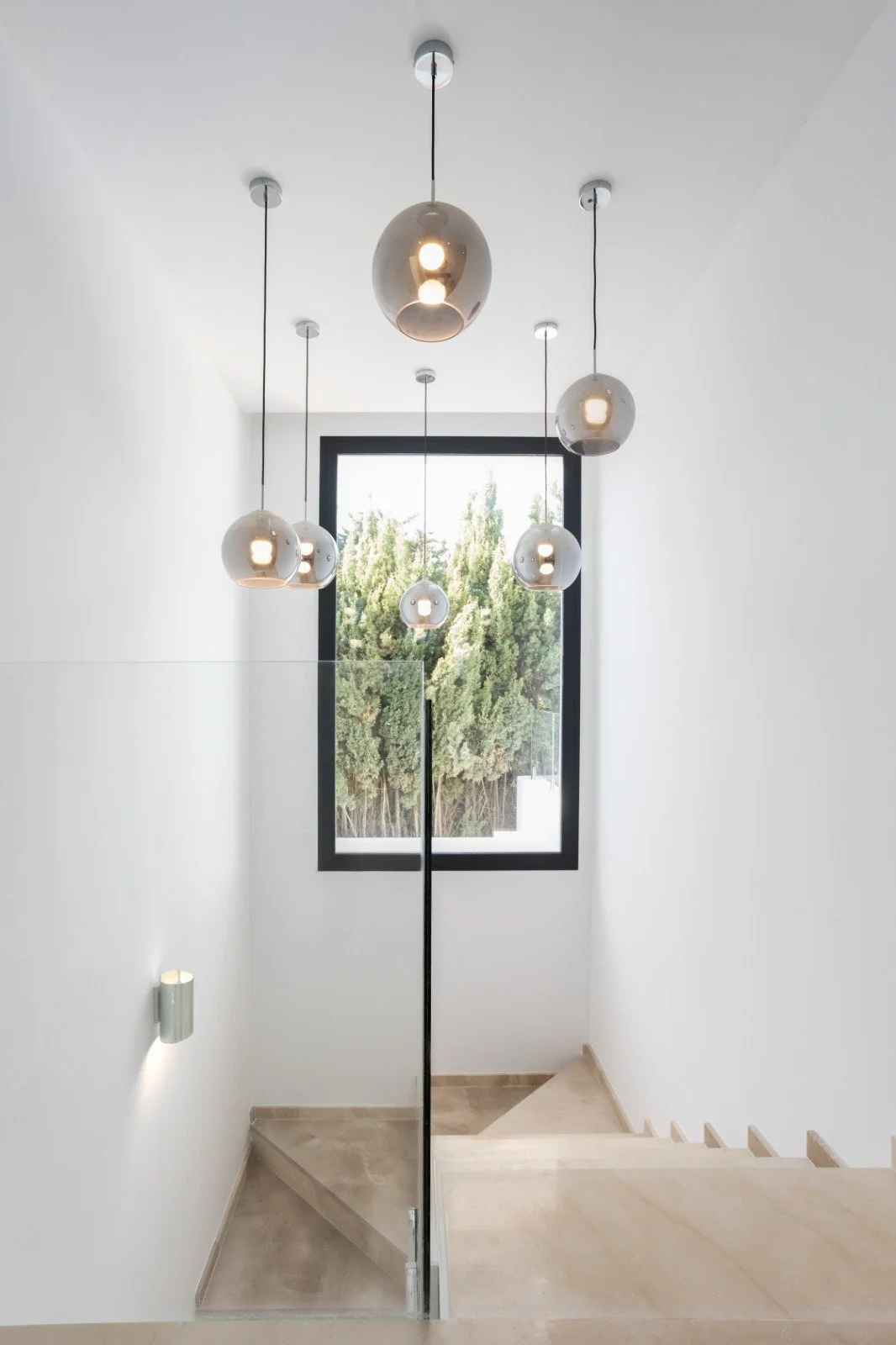 Modern staircase with white walls, a large window showing green trees, and a ceiling of hanging spherical pendant lights.