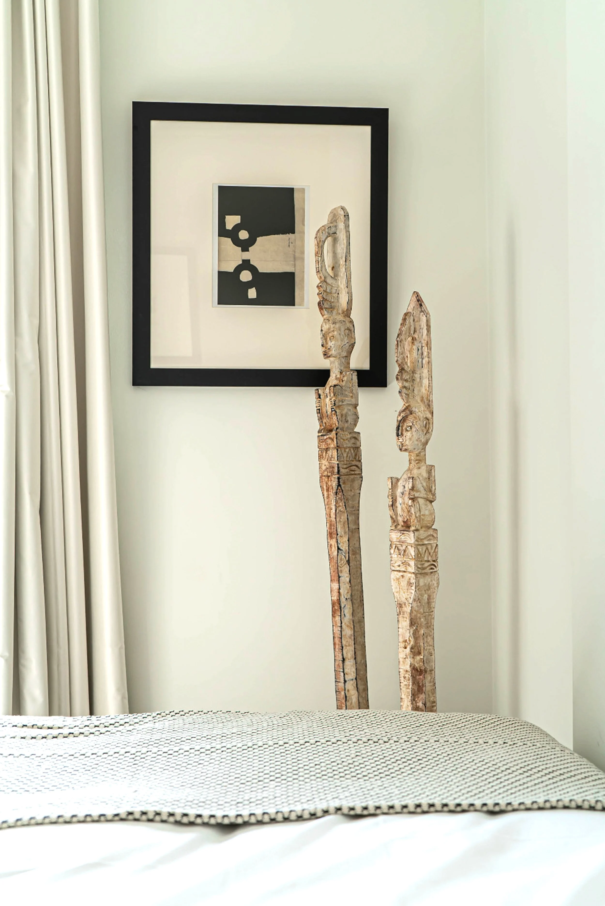A bedroom with two tall wooden carved tribal statues, a black framed abstract art piece on the wall, white curtains, and a bed with a patterned throw.