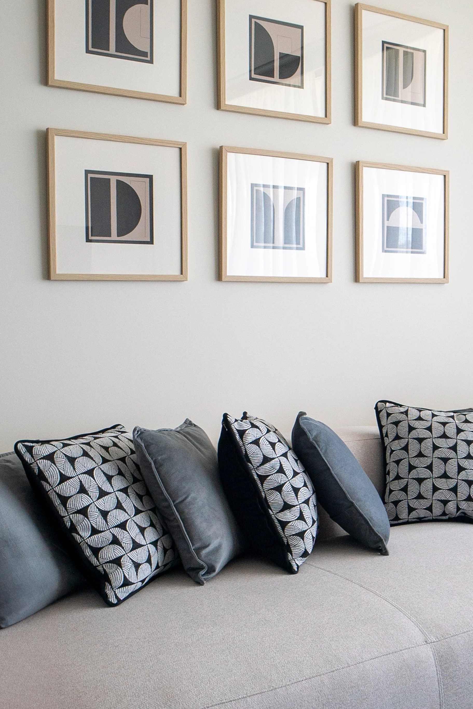 A collection of black and gray throw pillows with geometric and leaf patterns on a curved beige sofa, against a white wall decorated with six framed abstract geometric art pieces.