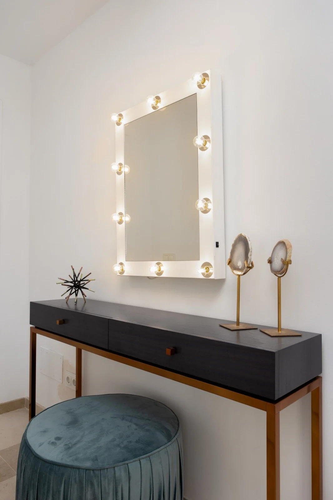 Interior entryway with vanity mirror surrounded by light bulbs, dark sideboard, decorative sculpture, and geode sculptures on stands, with a gray upholstered stool.