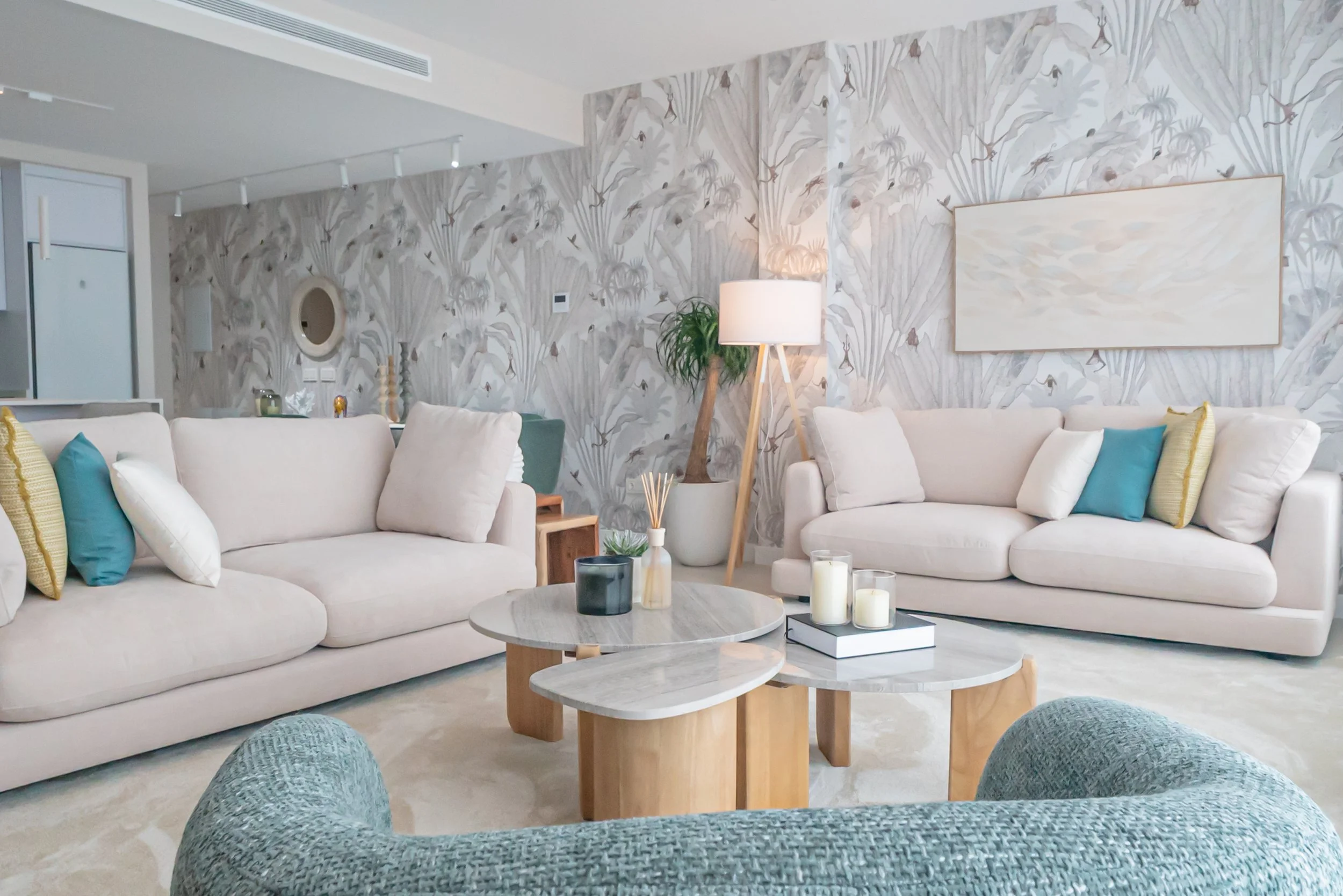 A cozy living room featuring wallpapera long beige sofa with pillows, a small armchair, a wooden coffee table, and a striped rug. There are large windows with white frames allowing natural light, and decorative vases on the left and in a wall niche.