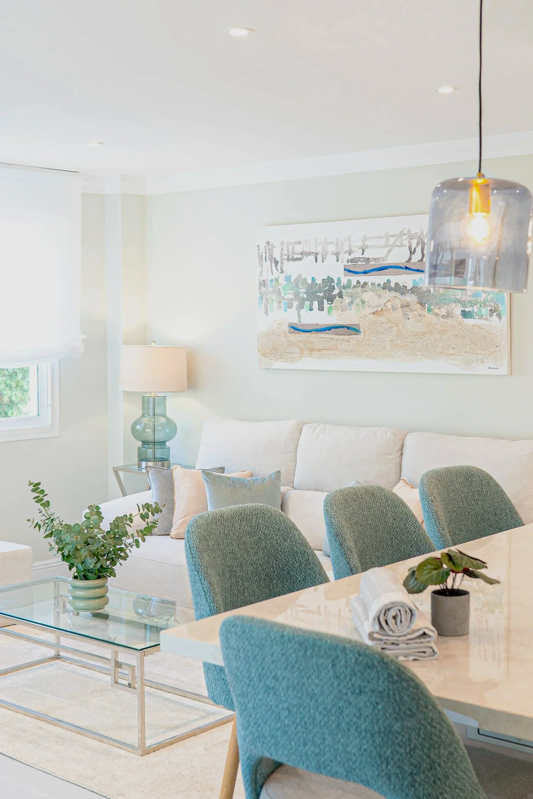Living room with a light-colored sofa, blue and beige throw pillows, a glass coffee table with a green plant and rolled towels, a side table with a glass lamp, a large abstract painting on the wall, several green upholstered chairs around a dining table, and a pendant light.