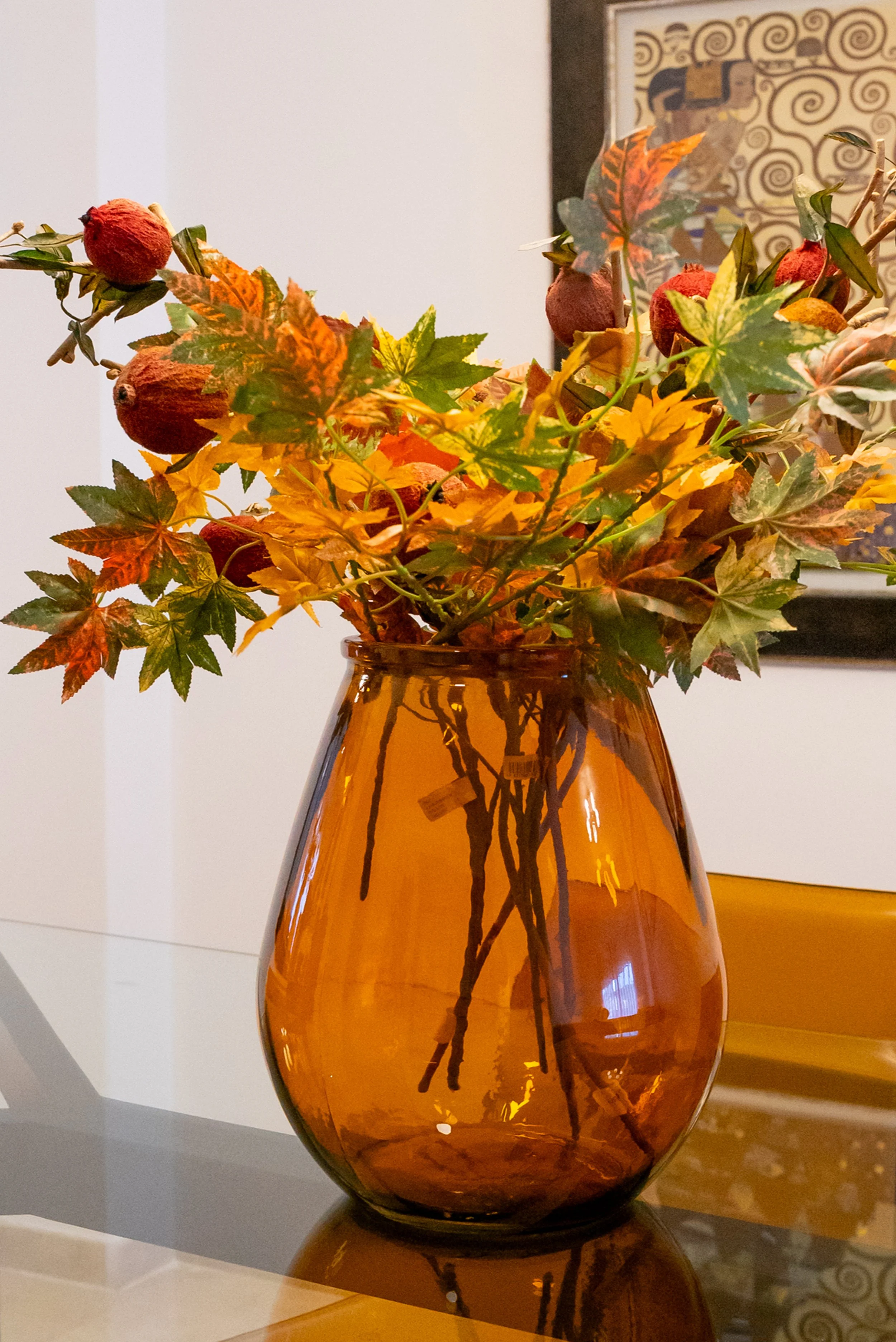 A brown glass vase containing autumn leaves and dried seed pods, placed on a reflective surface with a white wall and framed artwork in the background.