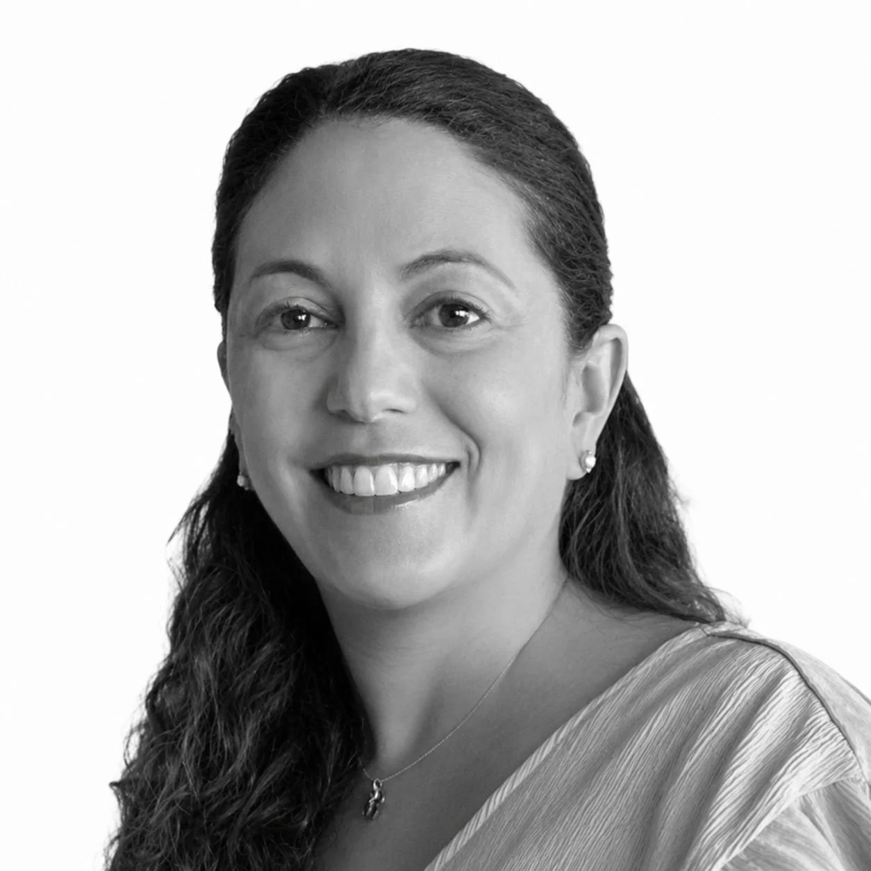 A smiling woman with long, wavy dark hair, wearing earrings, a necklace, and a striped top, facing slightly to the left against a plain background.