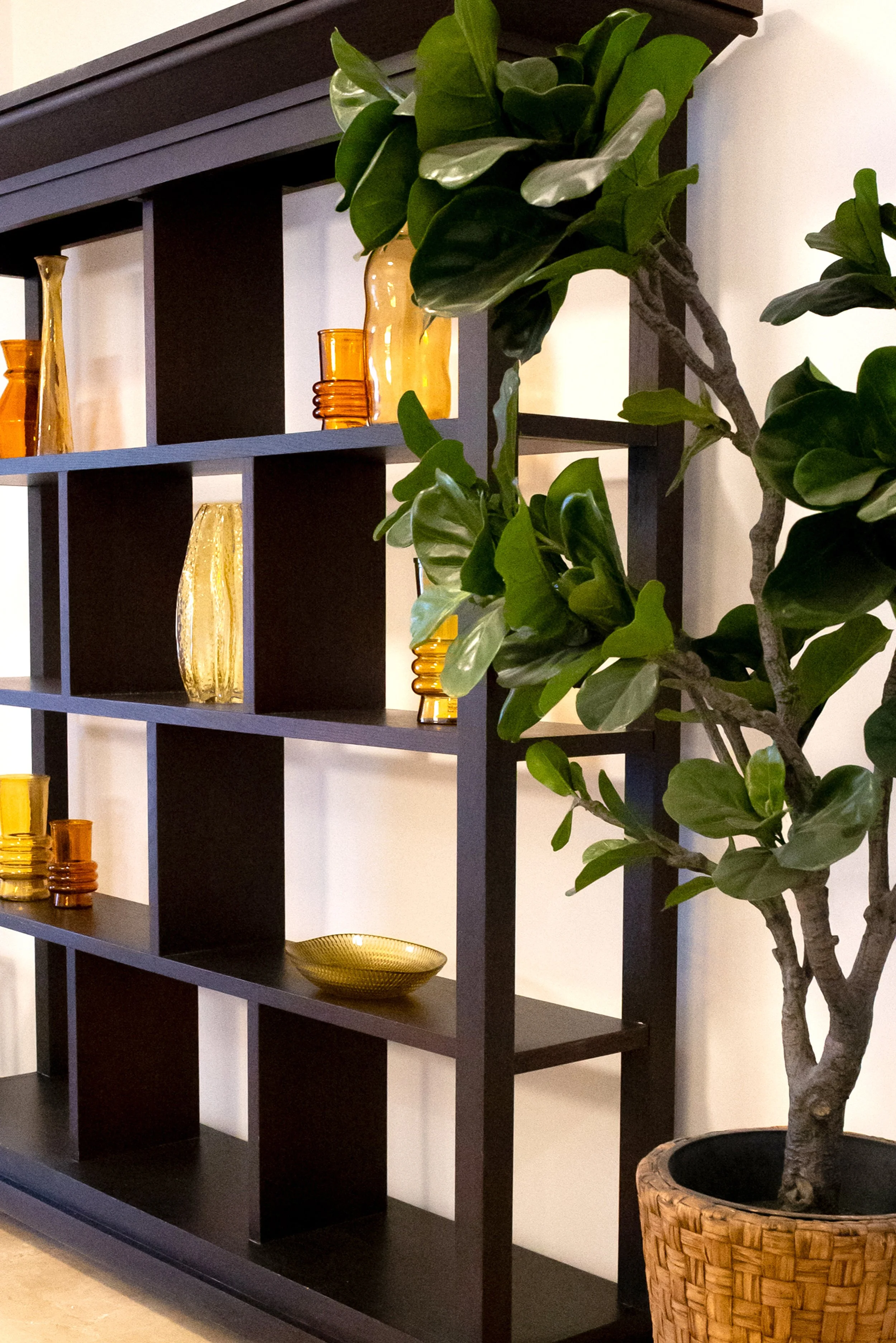 A black wood bookshelf with multiple compartments, decorated with amber-colored glass vases and bowls, and accompanied by a large potted plant with broad green leaves on the right.